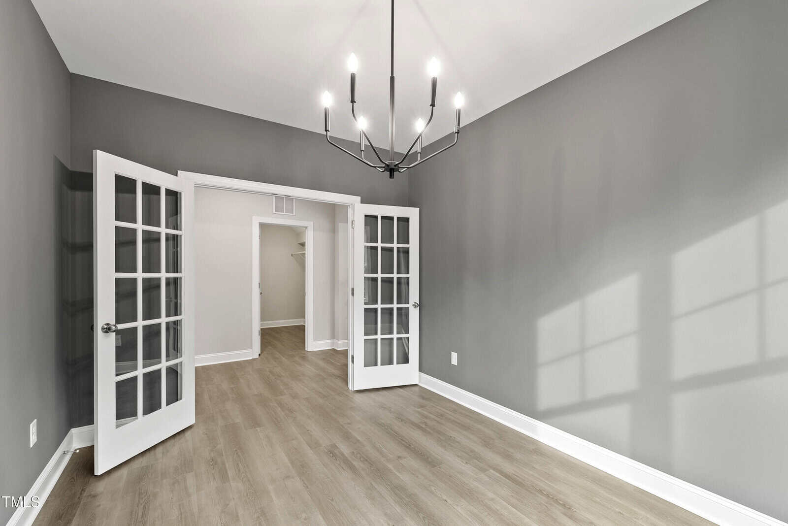 A bright, empty room with gray walls, light wood flooring, a modern black chandelier, and French doors with glass panels opening to a hallway and closet. Sunlight casts window shadows on the wall.