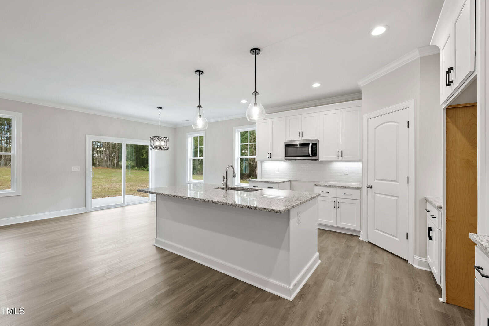 Modern kitchen with white cabinets, granite island, stainless steel appliances, pendant lights, wood flooring, and large windows overlooking a green backyard. Adjacent dining area with glass sliding doors.
