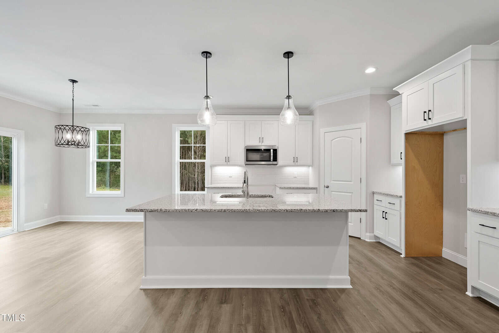 Modern kitchen with white cabinets, stainless steel appliances, a large granite island with two pendant lights overhead, and wood flooring. Large windows and a dining area are visible on the left side.