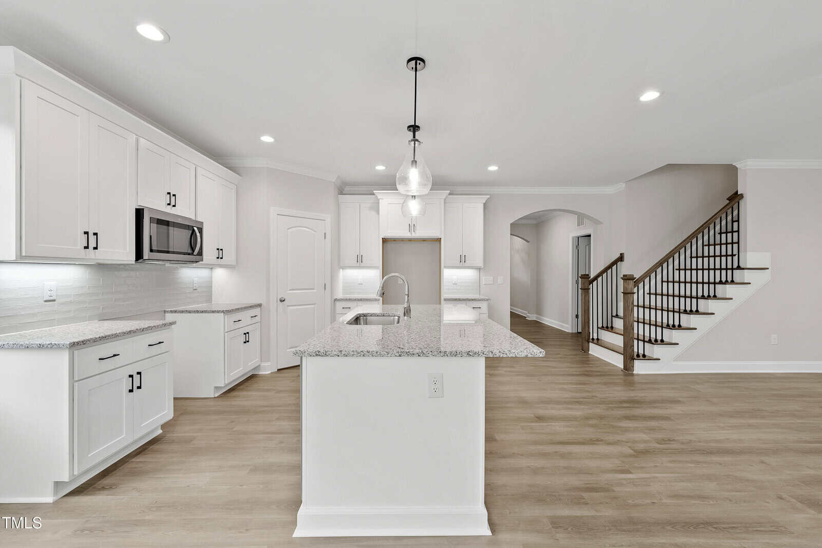 Modern kitchen with white cabinets, granite countertops, an island with a sink, stainless steel appliances, pendant lights, and wooden flooring. A staircase with dark railings is visible to the right.