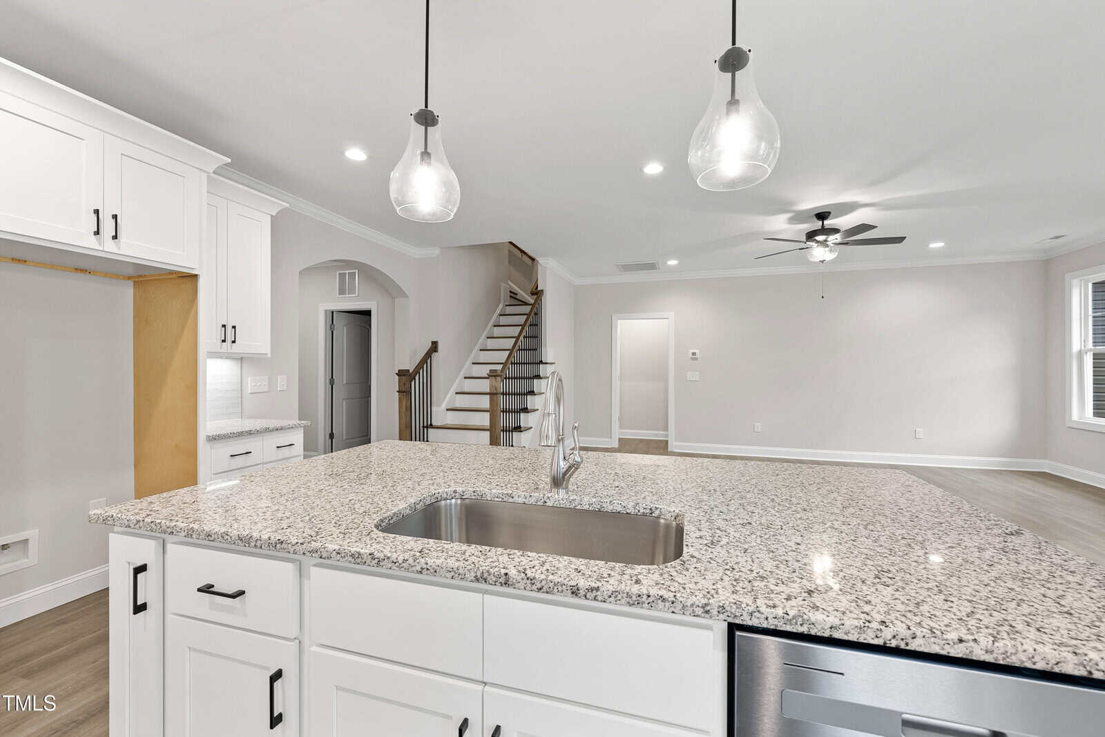 Modern kitchen with a granite island, sink, and stainless steel dishwasher, connected to an open living area with wood floors, white cabinets, and a staircase leading upstairs.