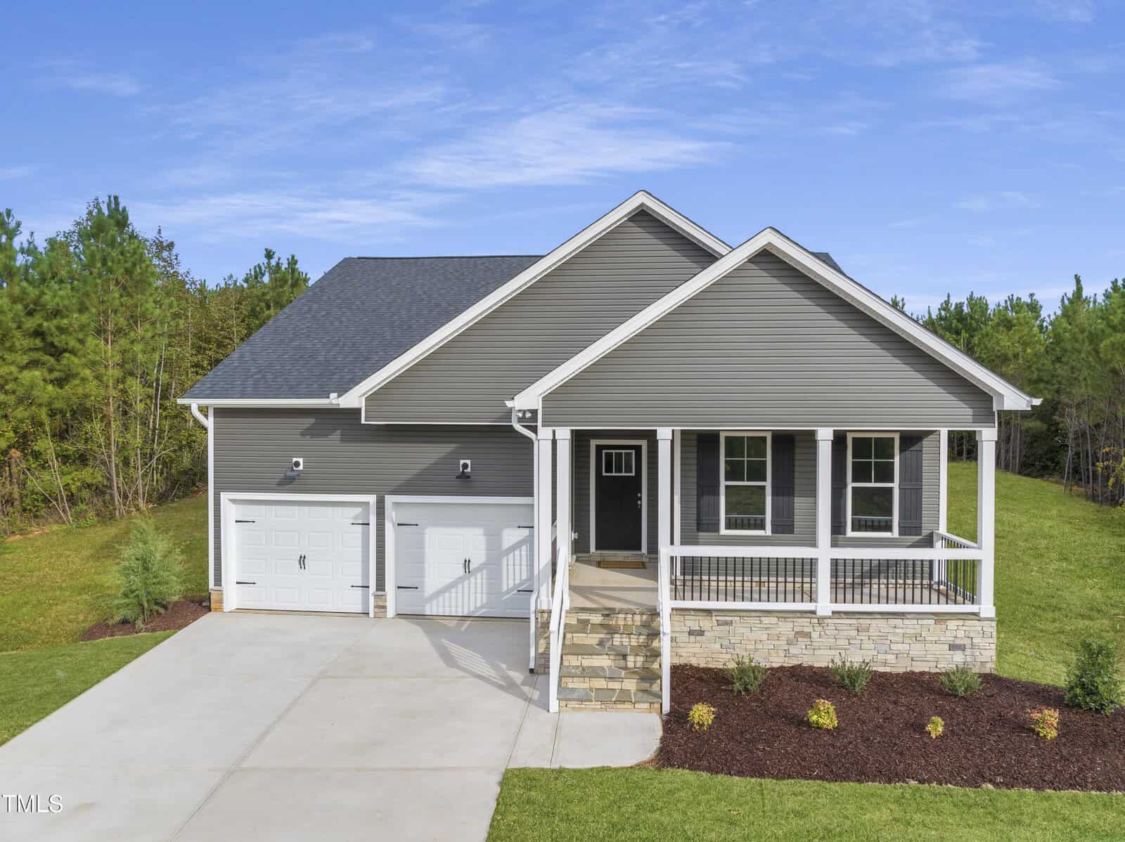 A modern single-story gray house with a covered front porch, white trim, and a double garage. The house sits on a neatly landscaped yard with green grass, shrubs, and a concrete driveway, surrounded by trees.