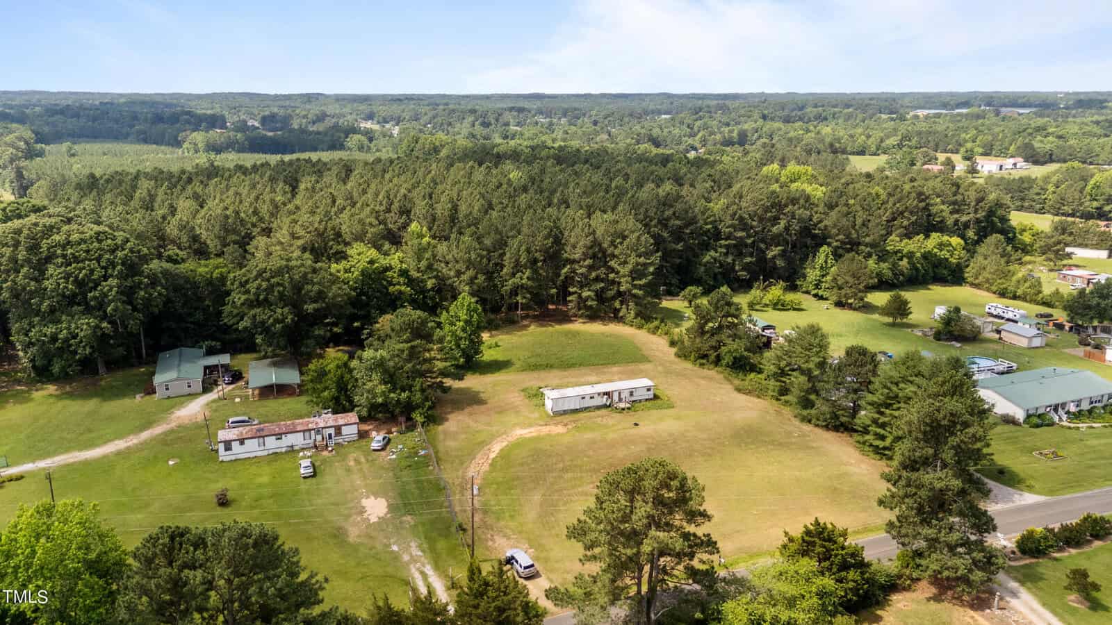 Aerial view of a rural area with multiple homes and trailers at 794 Club Pond Road, Henderson NC, surrounded by vast greenery and dense trees. A curved dirt road runs between the properties. The horizon shows distant forests and a slightly cloudy sky.