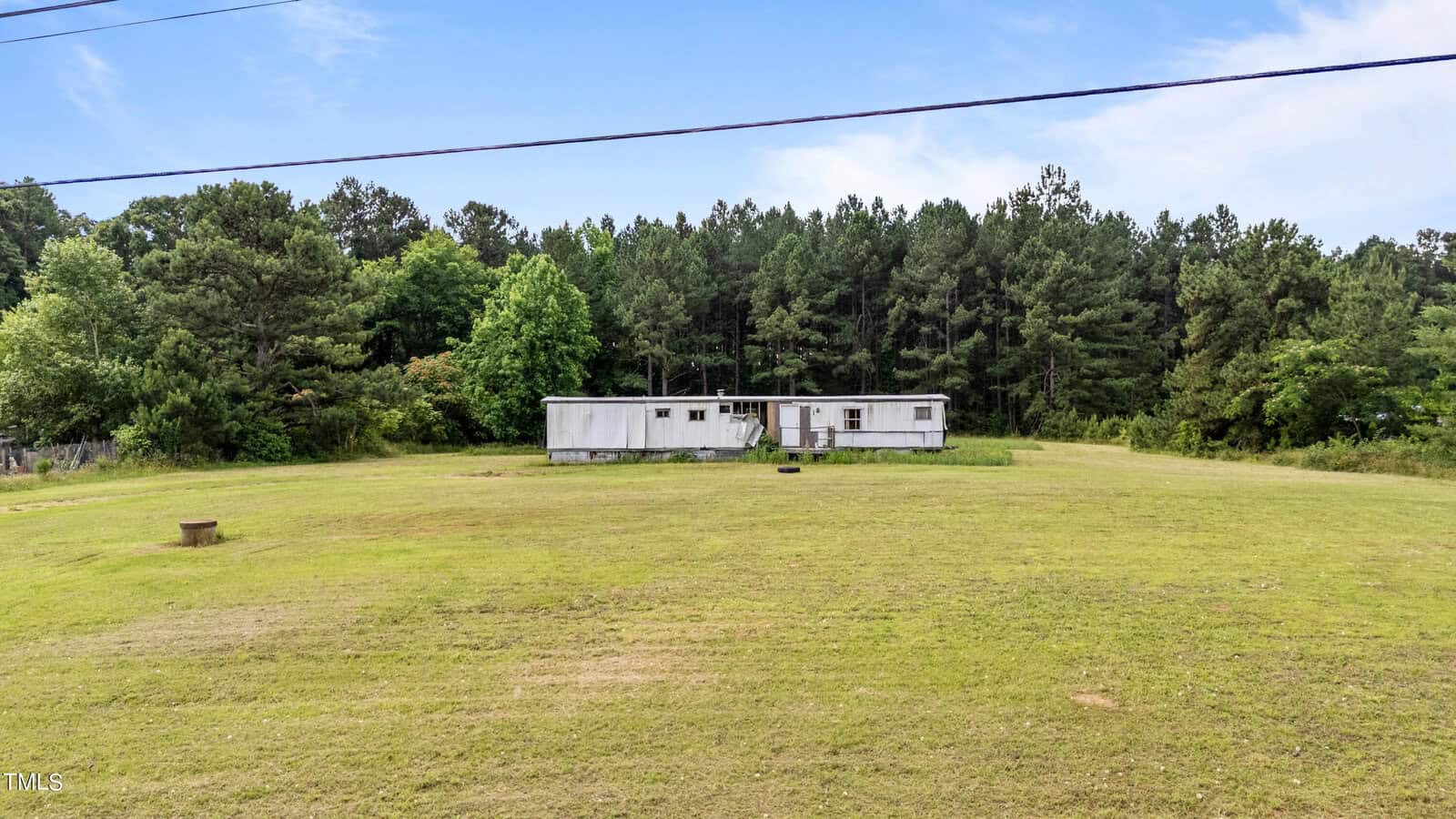 A long white trailer sits on a spacious, well-maintained grassy field with a backdrop of dense green trees under a partly cloudy sky. Power lines are visible at the top of the image.