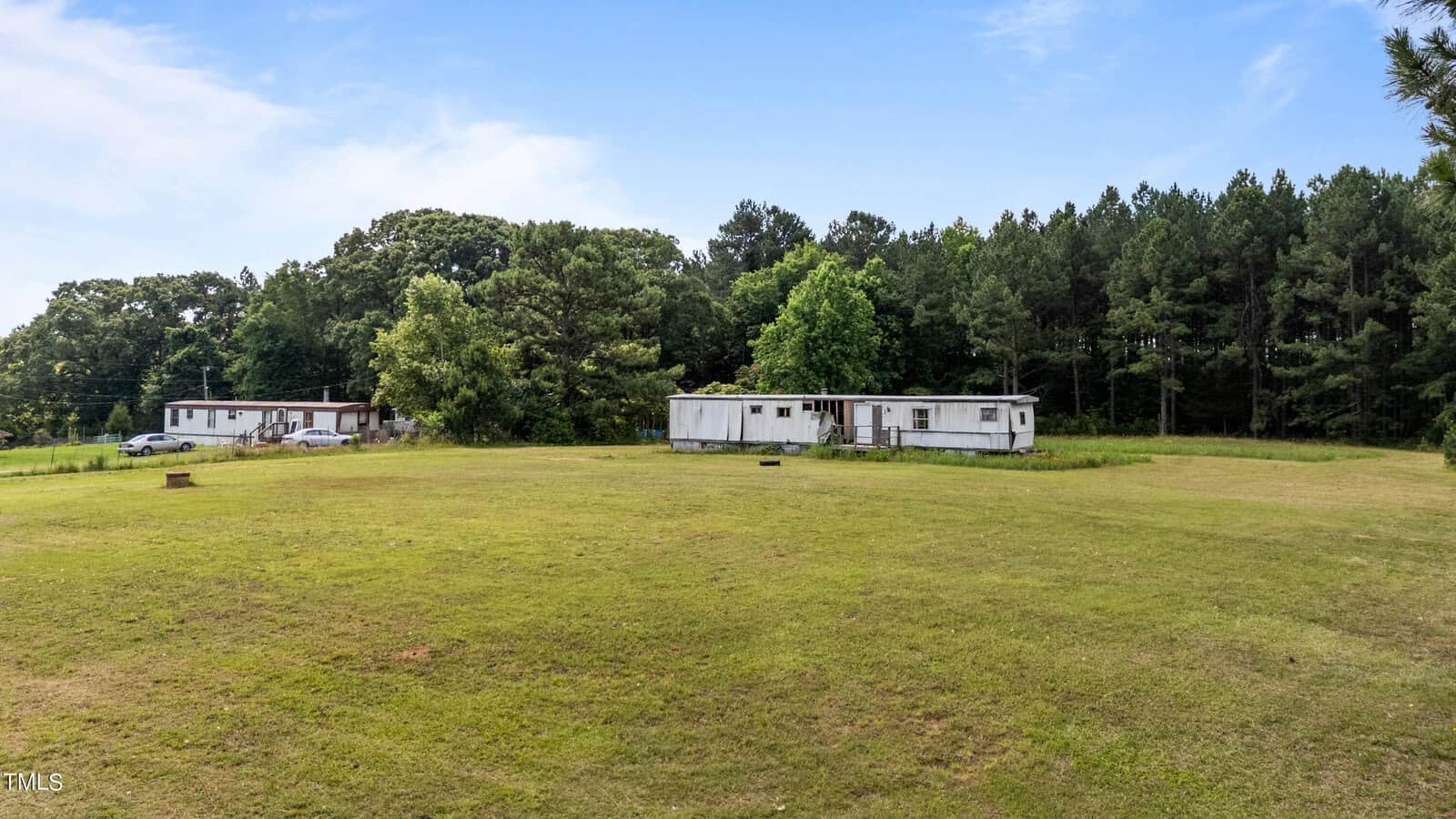 A grassy field with two white mobile homes set against a backdrop of dense, green trees under a bright blue sky with scattered clouds. One mobile home is visible on the left side and the other in the center. The terrain is flat and open.