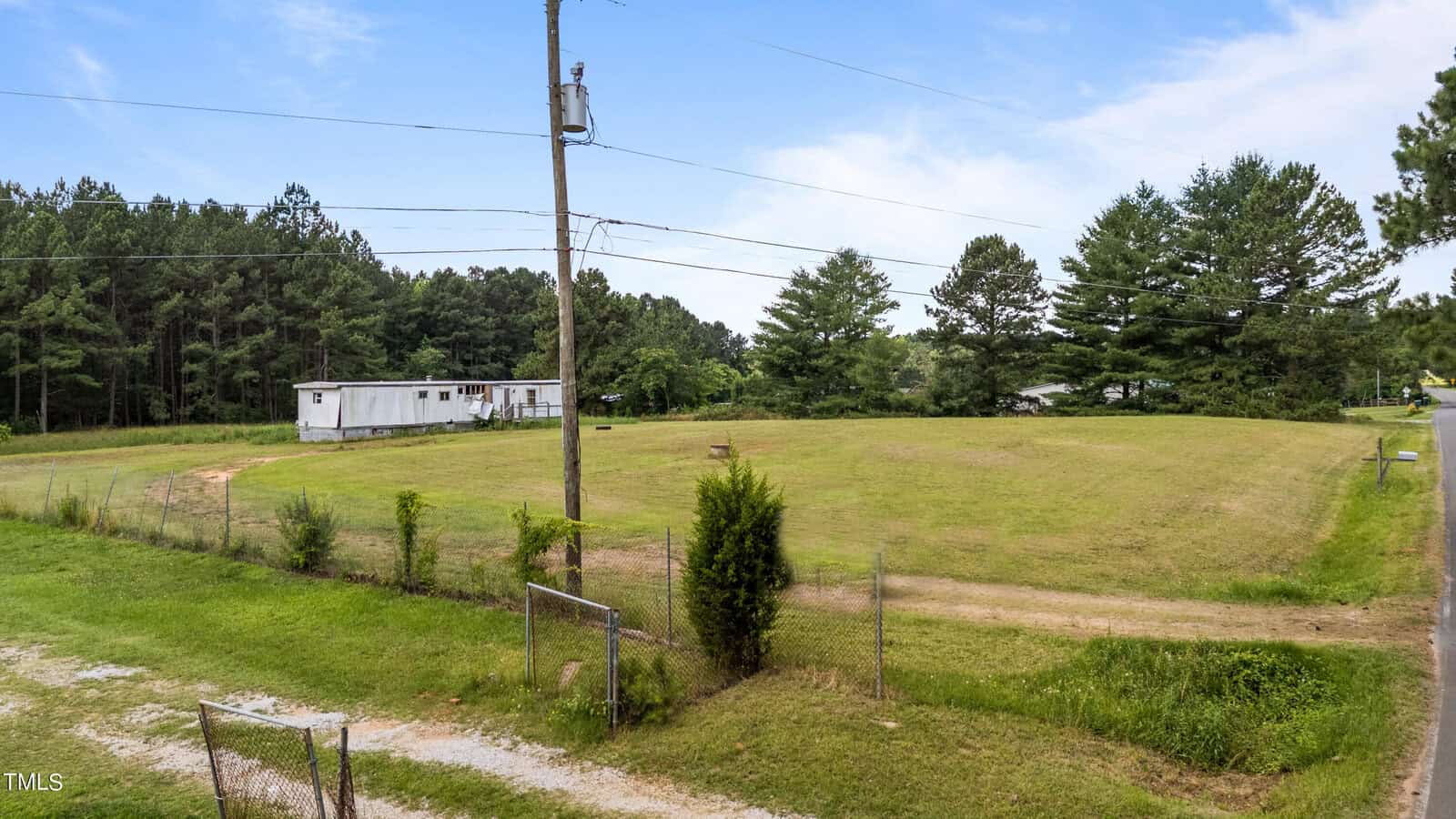 A wide view of a rural property with an open grassy field bordered by a fence. A white trailer sits in the distance near a dense forest of tall trees. Power lines cross the sky, and a dirt path leads towards the trailer. The sky is clear with a few wispy clouds.