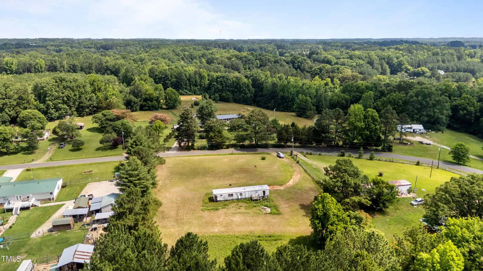 Aerial view of a rural area featuring a mobile home in a large, open, grassy field surrounded by trees. A road runs horizontally through the center of the image, with additional houses and structures visible along and near the road. Forested areas extend into the distance.