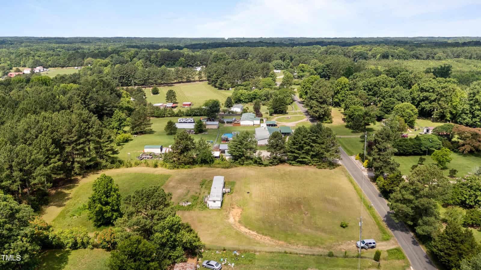Aerial view of a rural area featuring scattered houses, green fields, trees, and a winding road. The landscape is lush and predominantly green, with open spaces and a clear, blue sky in the background. A few structures and vehicles are visible.