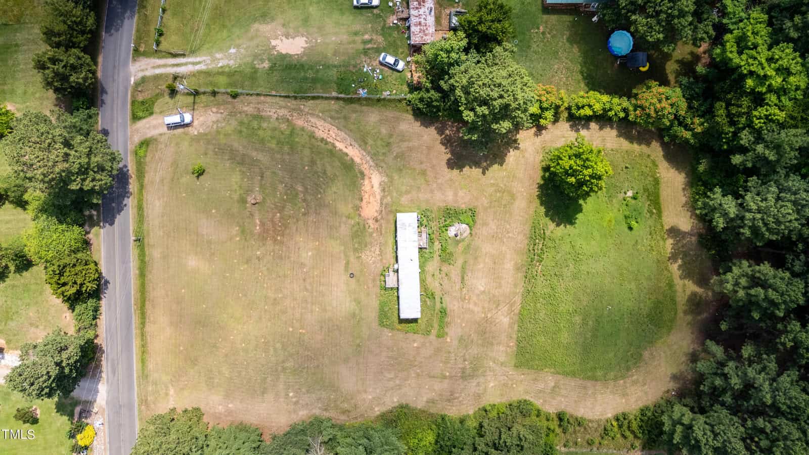 An aerial view of a grassy property featuring a long white building at the center. Surrounding areas include trees, a road on the left, and various parked vehicles. The landscape appears green and well-maintained, with separate sections of grass and sparse vegetation.
