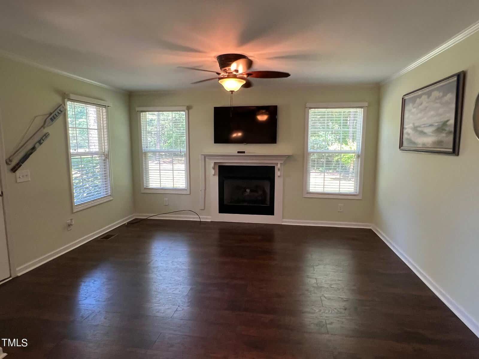 A well-lit living room with dark wood flooring, beige walls, and three large windows. There is a ceiling fan with a light fixture, a fireplace with a mounted TV above it, and a framed painting on the right wall. Large windows let in ample natural light.