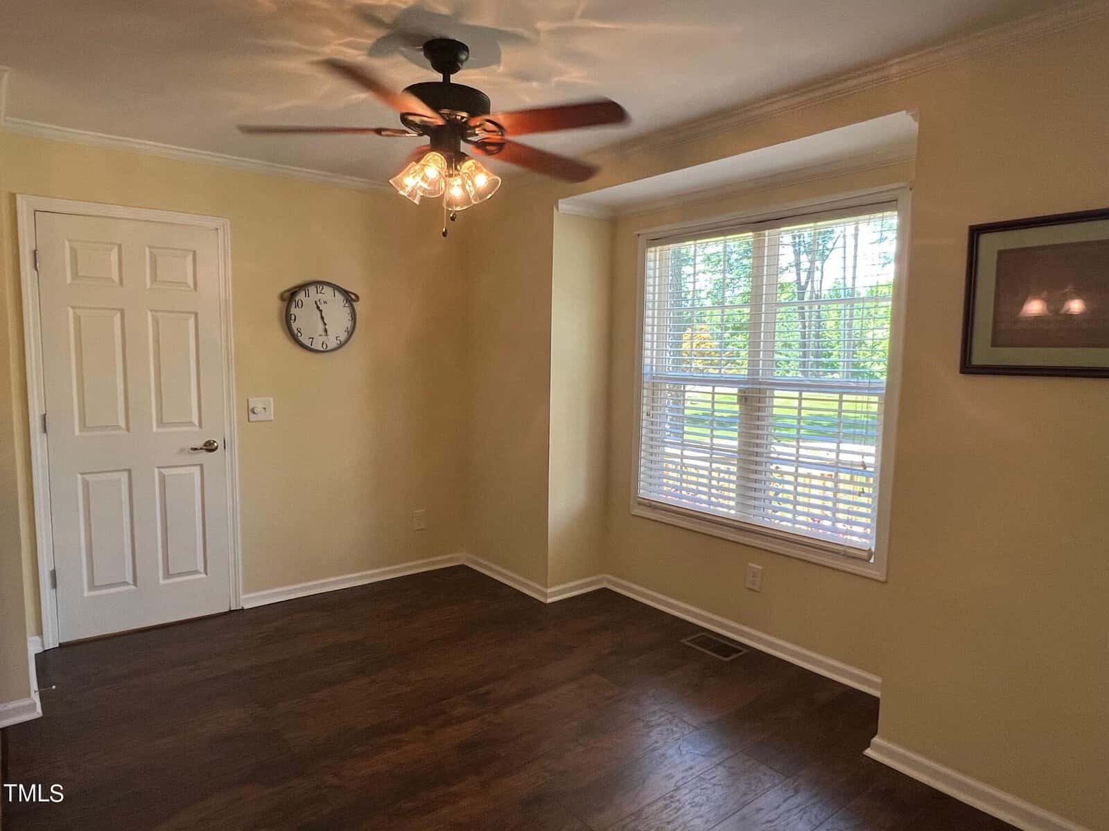 A small room with beige walls and dark wooden floors has a ceiling fan with lights, a white door, a wall clock, a framed picture, and a large window with blinds letting in natural light.