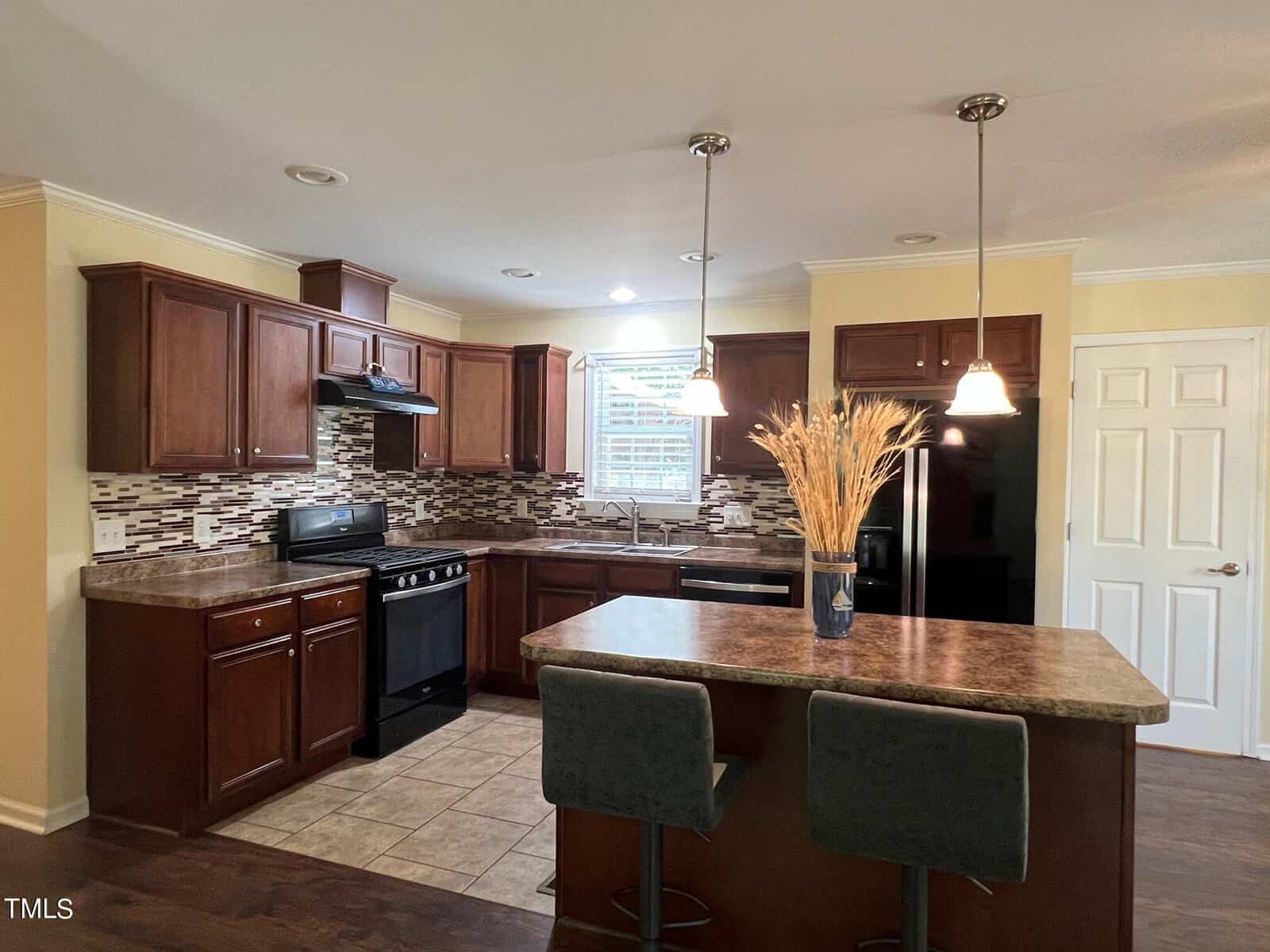 A modern kitchen featuring dark wooden cabinets, a tile backsplash, and stainless steel appliances. An island with a marble countertop has two bar stools and a vase with wheat stalks. Pendant lights hang above the island, and a window allows natural light to enter.