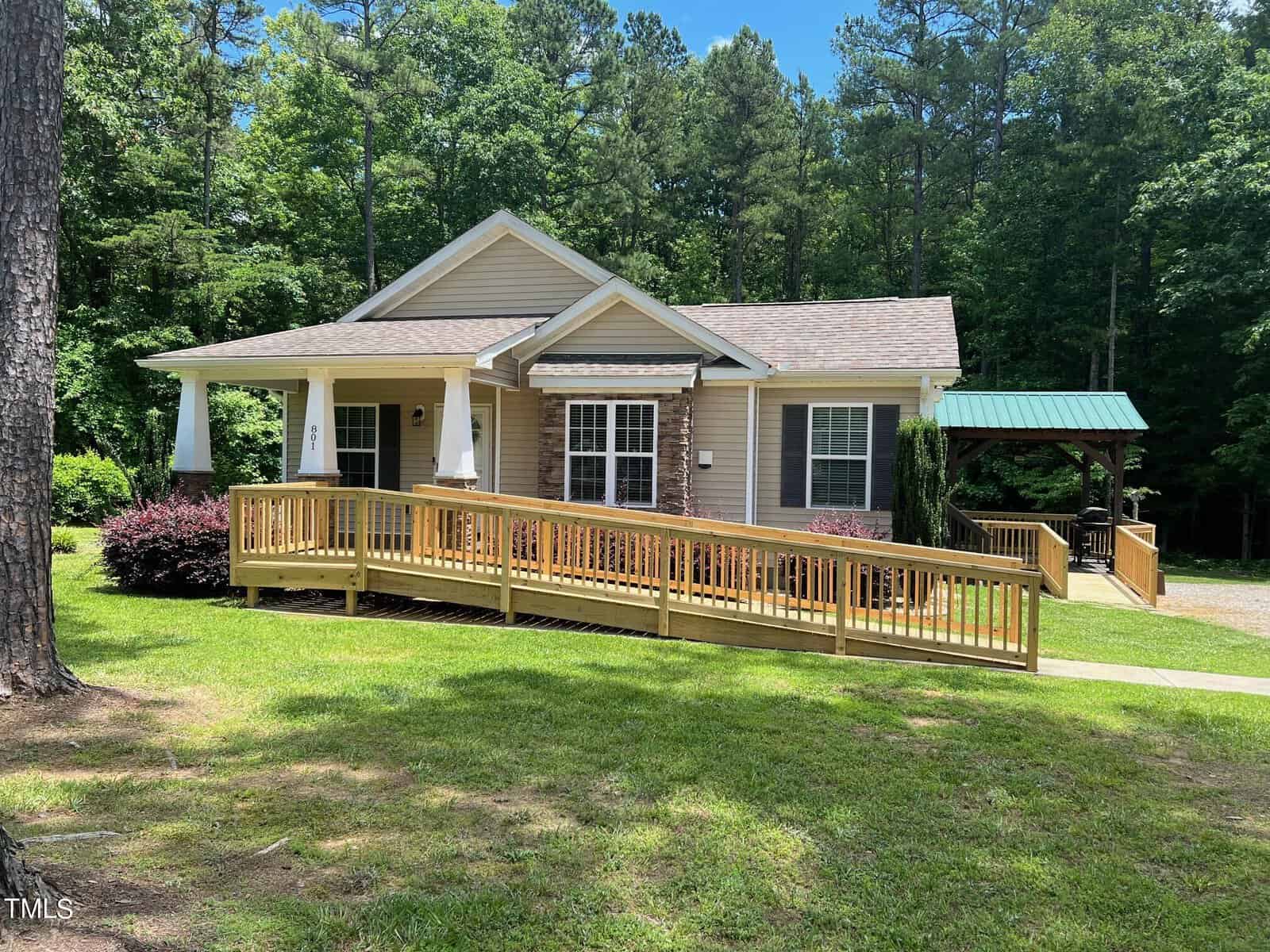 A small, single-story house with a beige exterior and a wooden wheelchair ramp leading to the front porch at 801 Port Drive in Henderson, NC. The house is surrounded by lush grass, trees, and bushes. A covered wooden gazebo is visible in the background under the clear, sunny sky.
