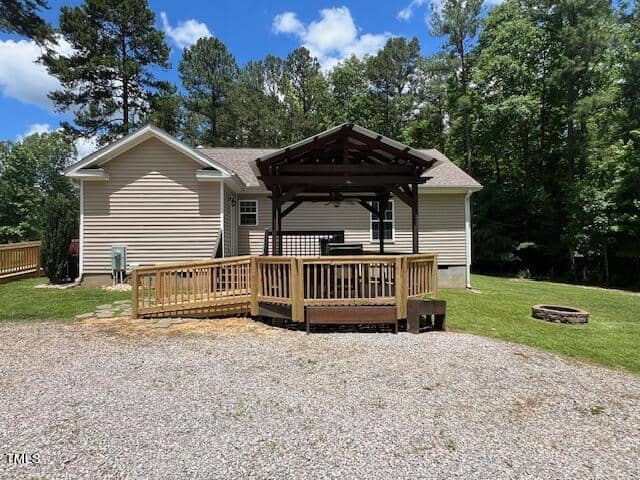 A light-colored single-story house with a small wooden deck and a pergola in the backyard. The backyard includes a gravel area, a grassy lawn, and tall trees in the background. Skies are blue with scattered clouds.