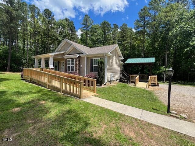 Single-story house with beige siding, situated in a lush, wooded area. The house has a large front porch with wooden railings and an accessibility ramp. A gravel driveway leads to a carport with a green roof. The sky is blue with scattered clouds.