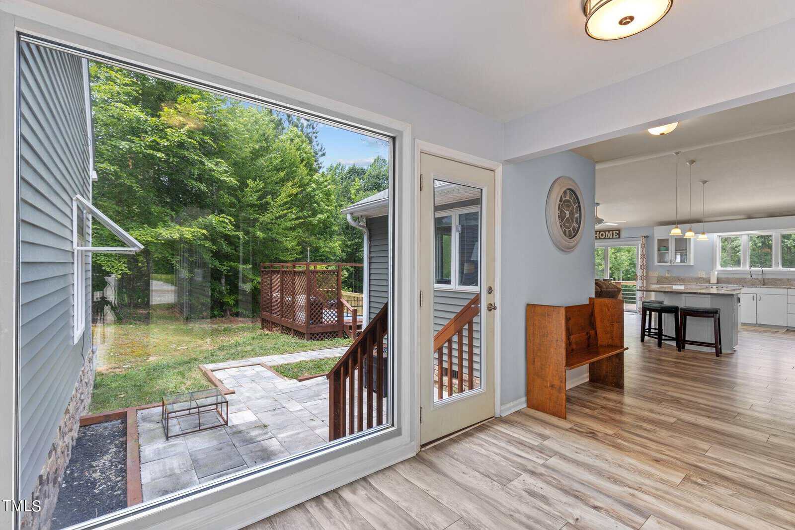 A bright interior features a large window and glass door overlooking a patio and greenery. Inside, light wooden floors lead to an open kitchen with white cabinets and modern lighting. A small bench sits near the doorway.