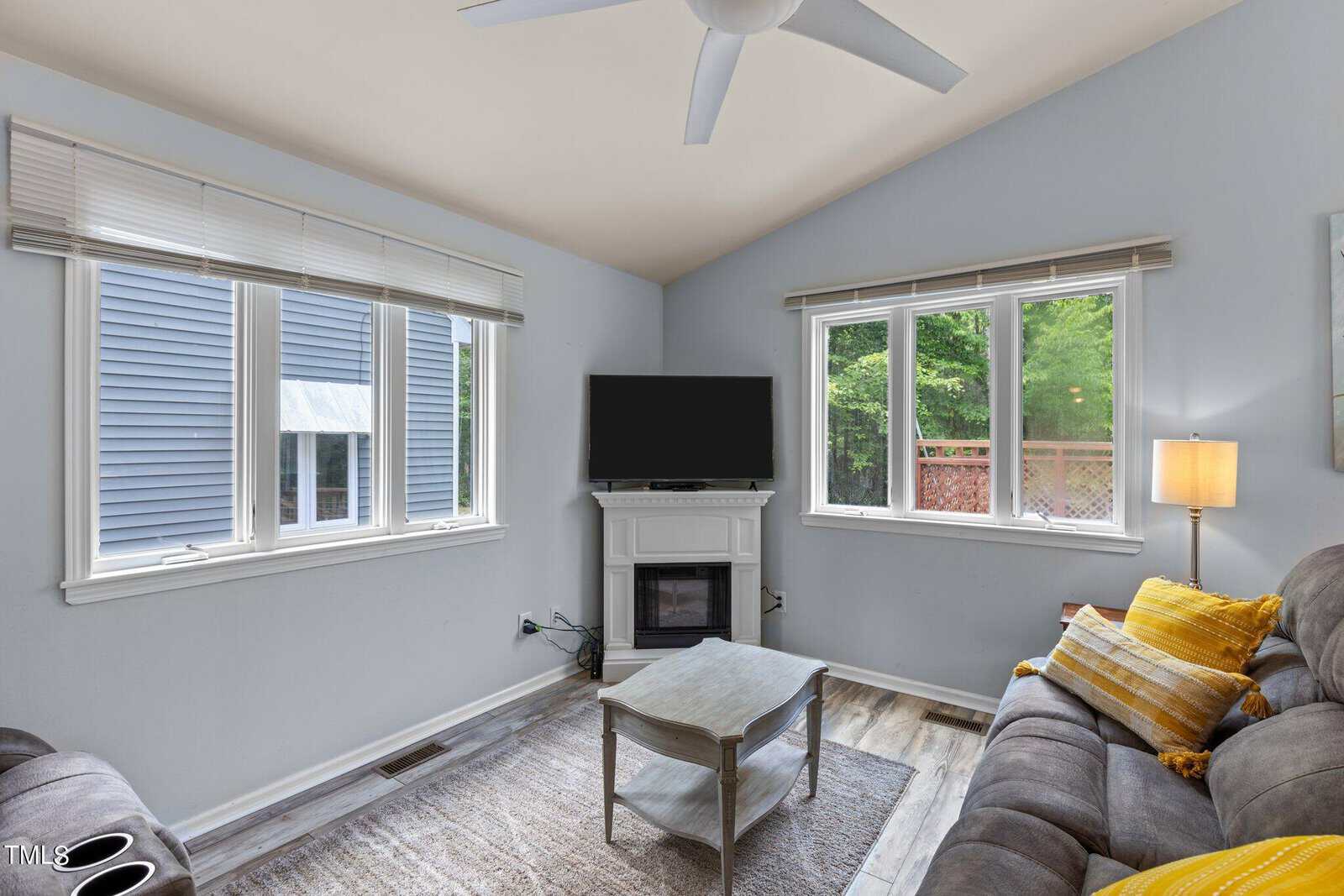 A cozy living room with pale blue walls, a sofa with yellow cushions, a white fireplace with a TV on top, and a wooden coffee table on a gray rug. Large windows let in natural light, revealing greenery outside. A ceiling fan hangs from the ceiling.