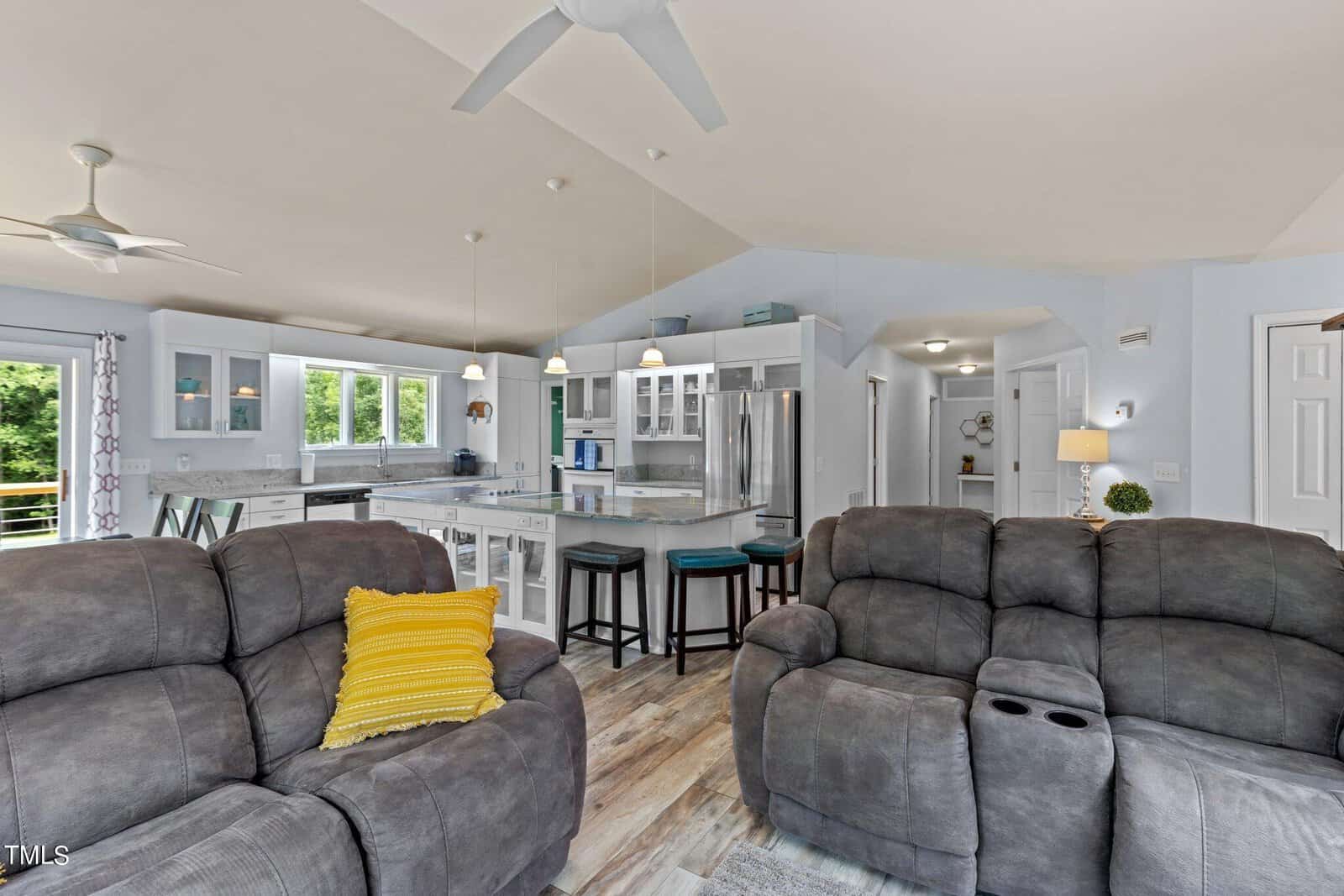 A modern living room and kitchen with gray sofas, a yellow cushion, pendant lights, and a high ceiling. The kitchen features white cabinets, a central island with stools, and natural light from windows.