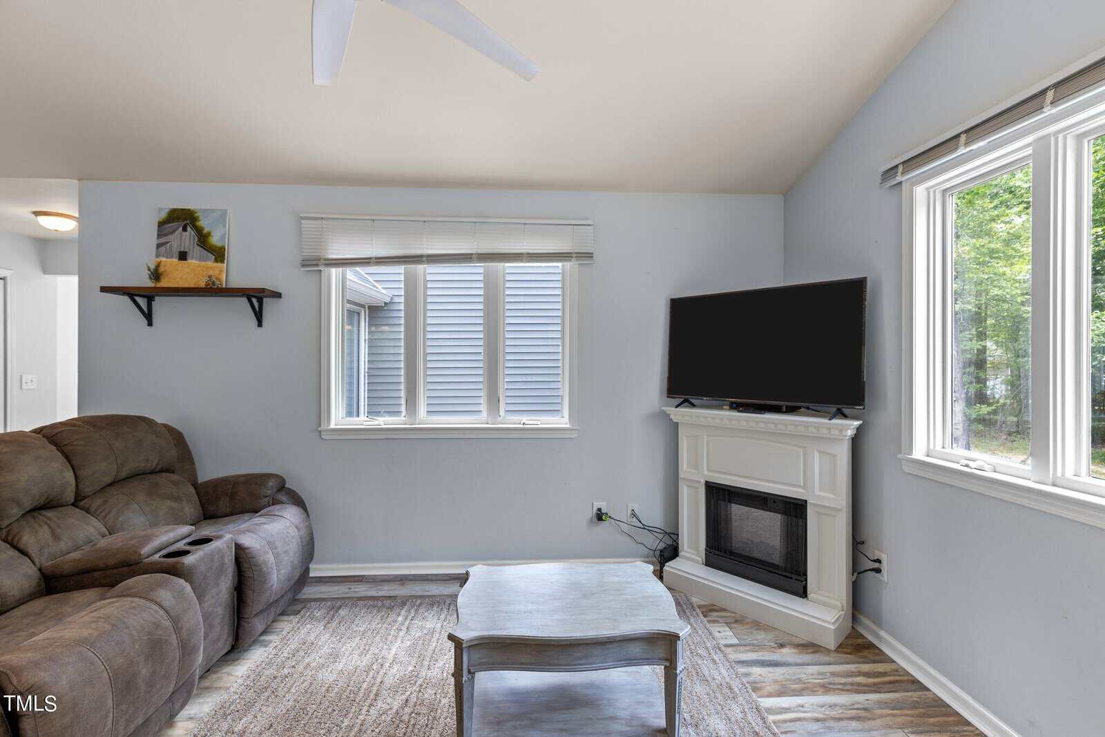 A cozy living room with a brown reclining sofa, a wooden coffee table, and a wall-mounted TV above an electric fireplace. The room has light blue walls, a high ceiling with a fan, and large windows letting in natural light.