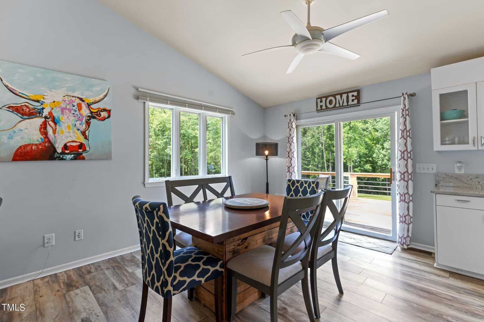 Dining room with a wooden table and chairs, large windows, and a sliding glass door leading to a deck. Walls are light blue with a colorful cow painting and a "HOME" sign above curtains. Ceiling fan overhead.