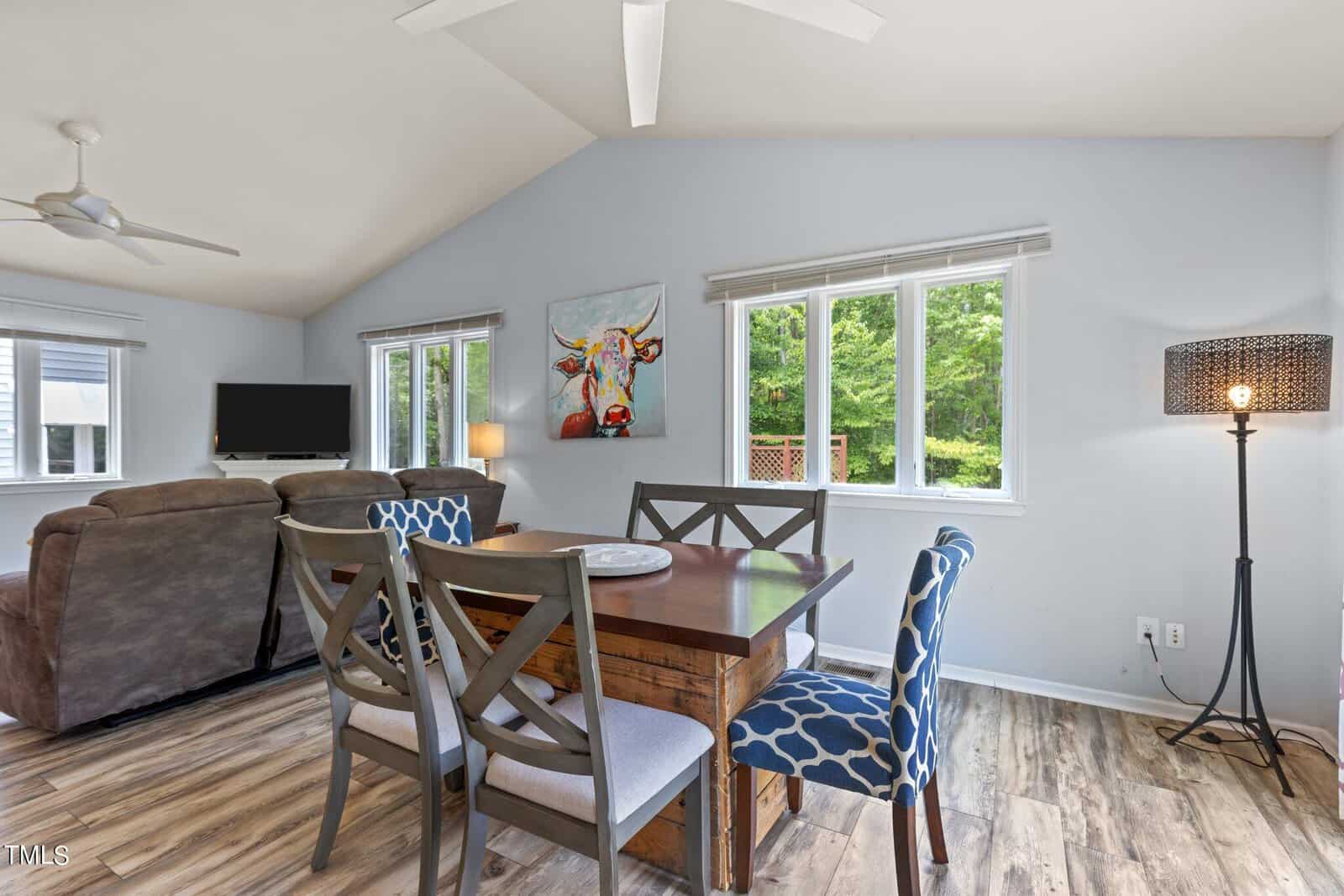 A bright living and dining area with light blue walls and large windows. There's a wooden dining table with patterned chairs, a cozy seating area with a TV, and a colorful cow painting on the wall. The floor is wood with a modern standing lamp.