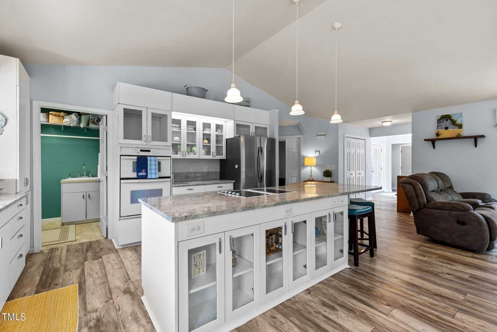 A modern kitchen with white cabinetry, an island with a marble countertop, and stainless steel refrigerator. Pendant lights hang above the island, and a cozy living area with recliners is visible in the background.