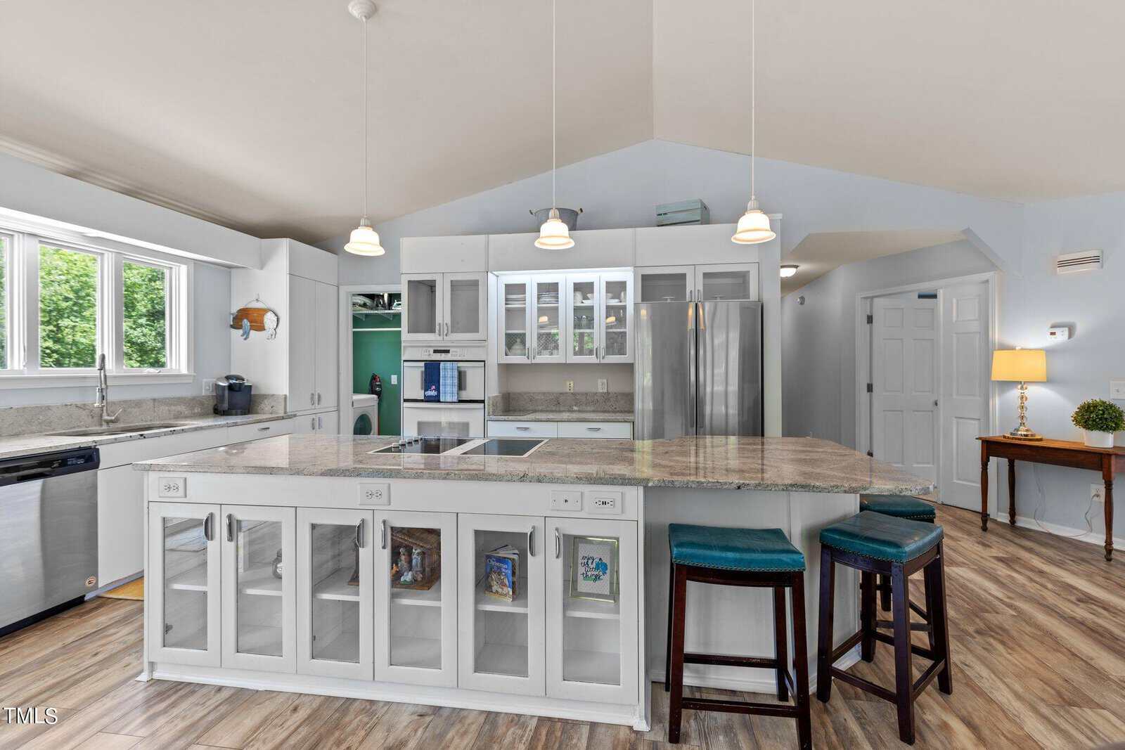 A spacious kitchen with white cabinets and a large island featuring glass-front storage and bar stools. Stainless steel appliances and pendant lighting add a modern touch. A window provides natural light, highlighting the wooden floor and light blue walls.