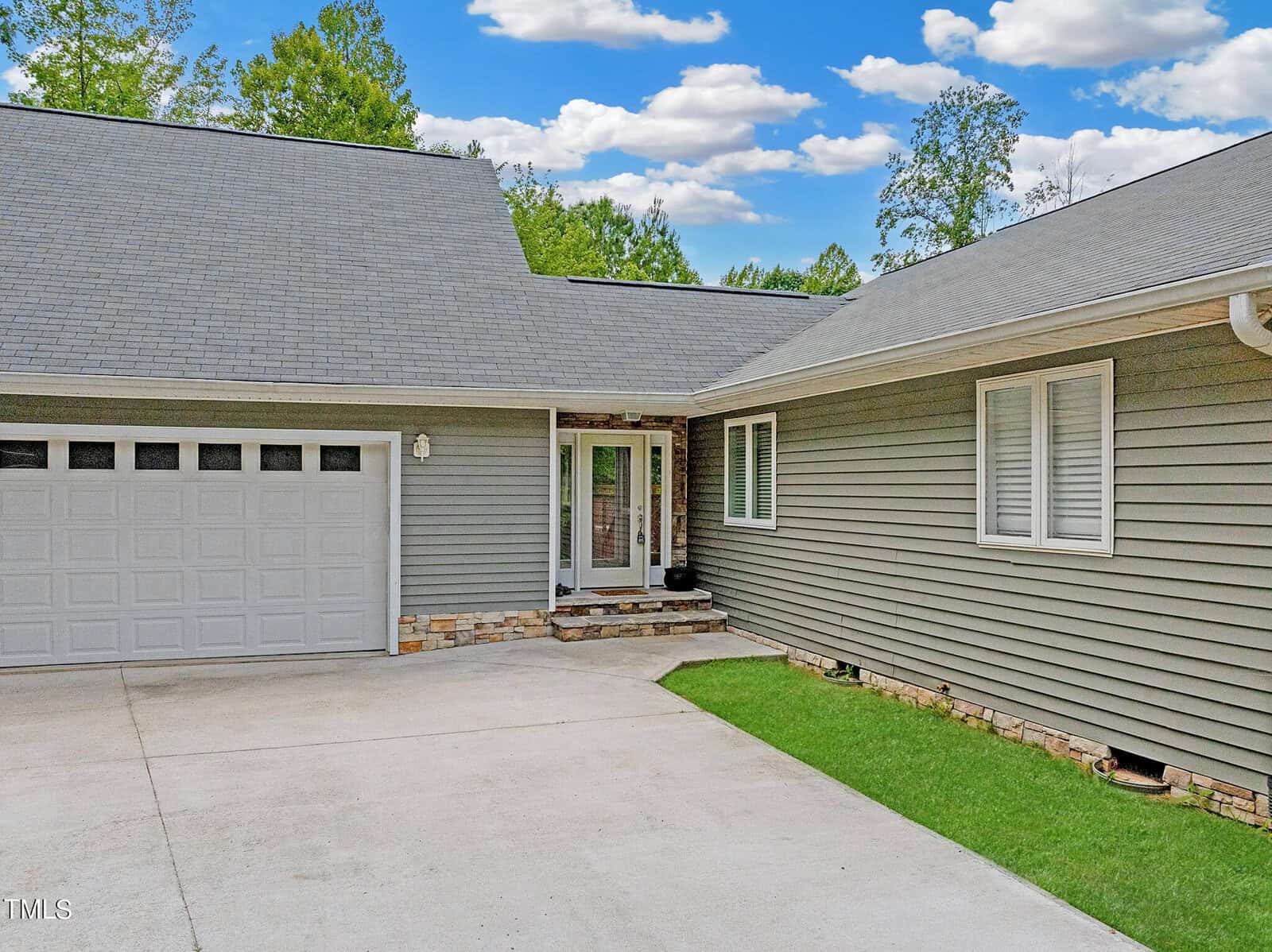 A modern gray house with a large garage and neatly trimmed lawn. The front entrance features a glass door with stone accents on the facade, under a clear blue sky with scattered clouds.