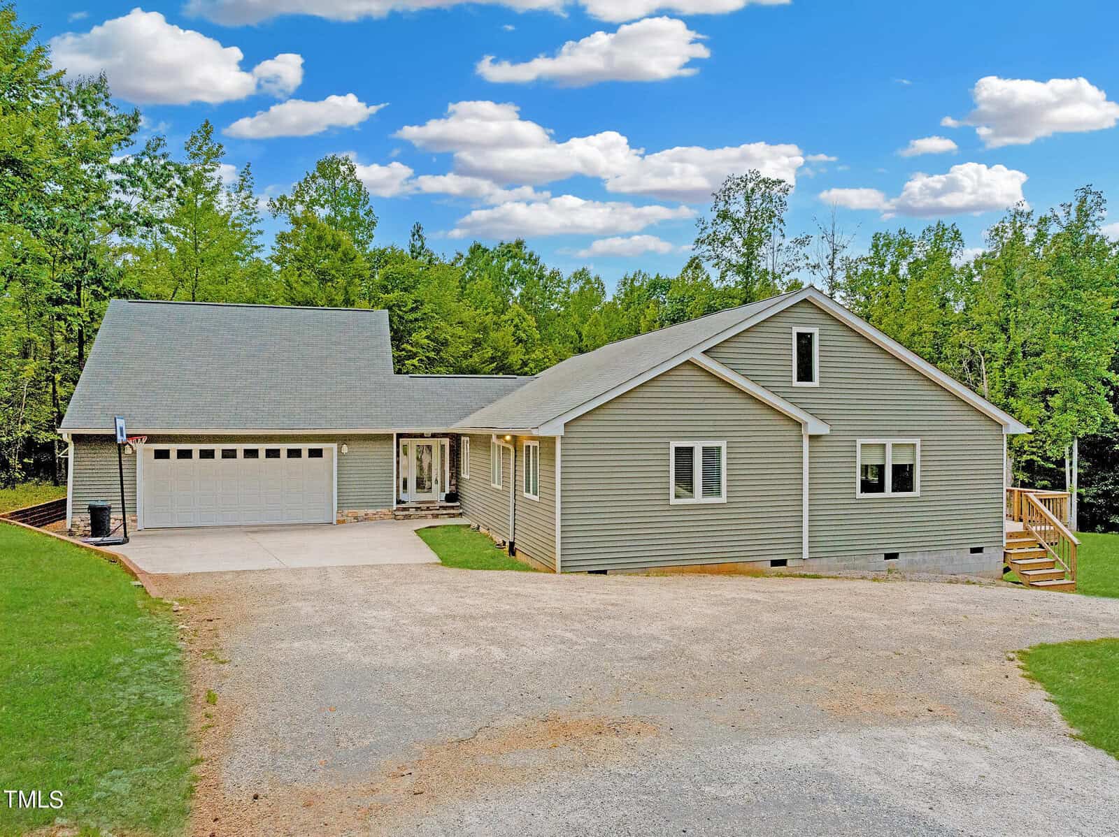 A single-story house with gray siding, located at 1060 Pebblebrook Drive in Wake Forest, features a two-car garage and a charming front porch. Surrounded by trees under a blue sky with scattered clouds, its gravel driveway and partially visible lawn complete the serene setting.