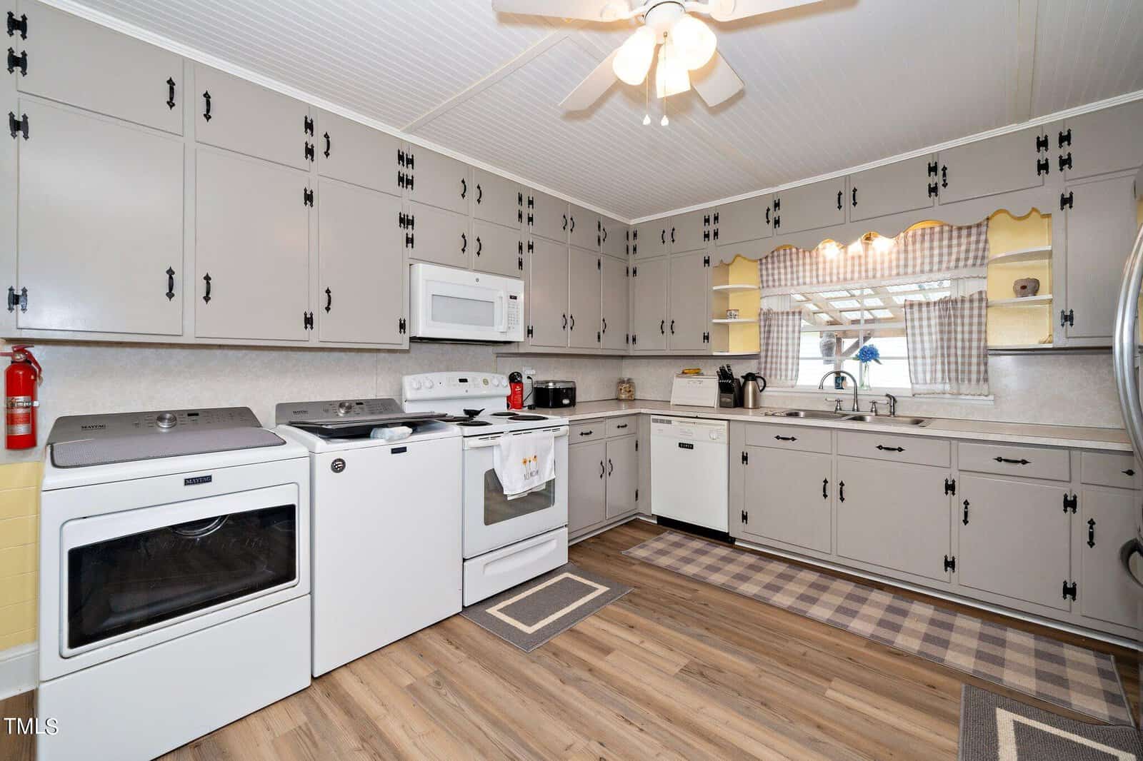 A vintage-style kitchen with gray cabinets, a white stove, microwave, and dishwasher. Wooden flooring and a ceiling fan complement the space. The window is decorated with checkered curtains, and a fire extinguisher is mounted on the wall.