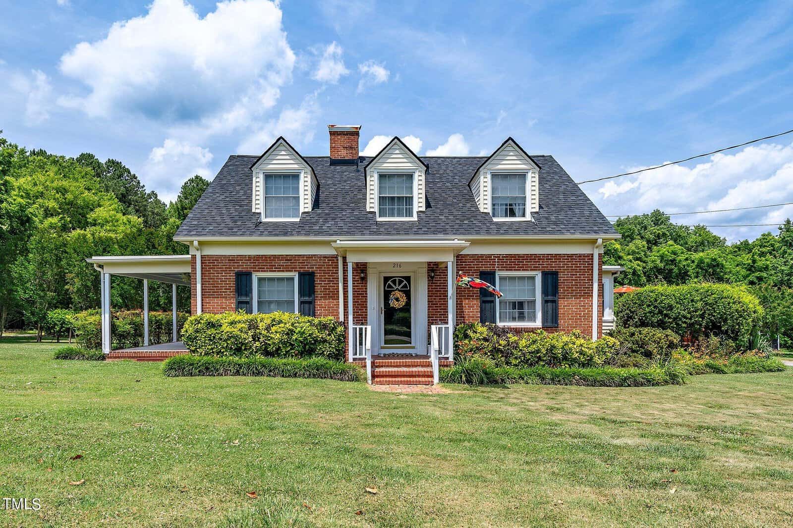 A charming red-brick house with white trim and a steep, dark gray roof sits at 216 Grove Street in Oxford. It features dormer windows, a white porch, and a well-maintained lawn and garden. A flag hangs near the entrance, surrounded by trees under a partly cloudy sky.