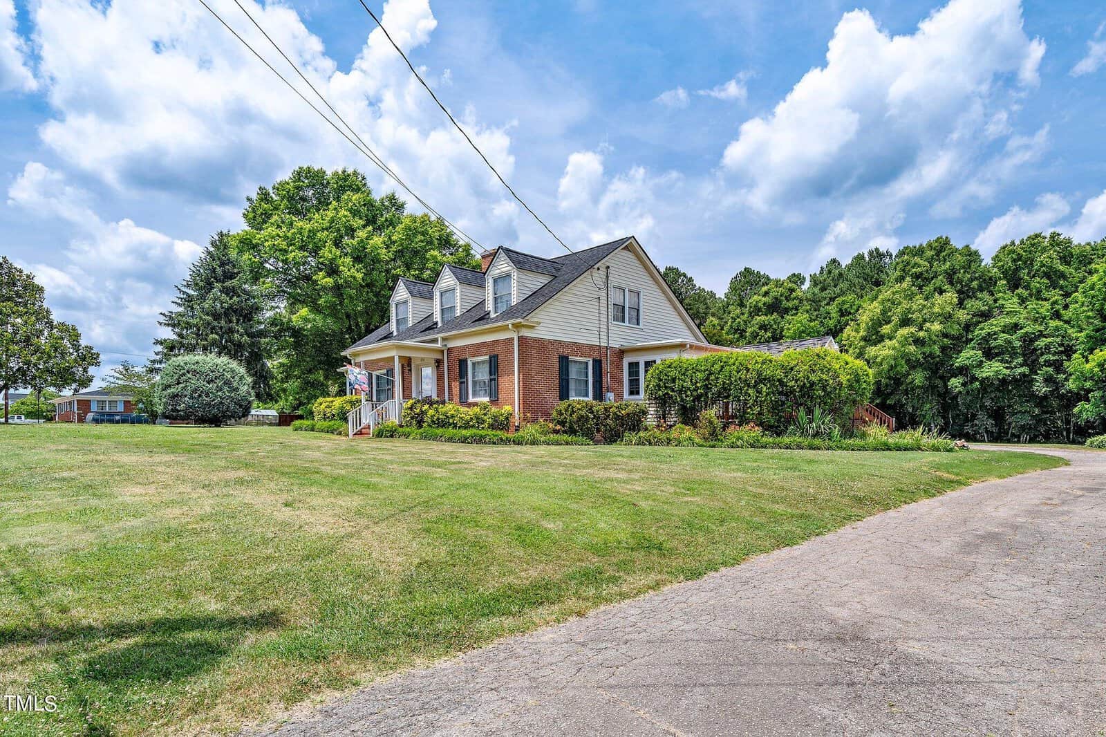 A brick house with a gabled roof and white dormer windows sits on a large green lawn. A paved driveway curves towards the house, and lush trees surround the property under a partly cloudy sky.