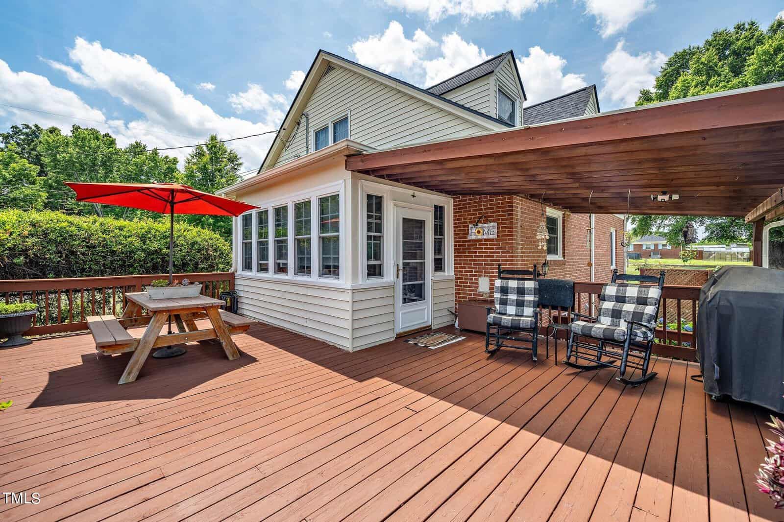 A wooden deck attached to a house features a picnic table with a red umbrella, two chairs with checkered cushions, and a barbecue grill. Lush greenery surrounds the deck under a partly cloudy sky.