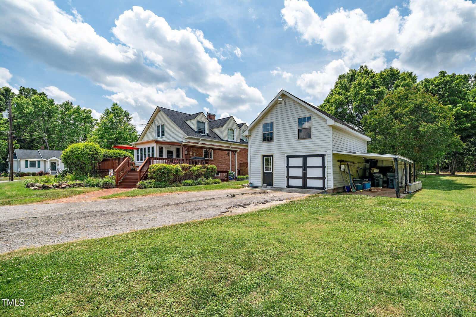 A two-story house with a wraparound porch is nestled in a grassy area under a partly cloudy sky. A white garage with an attached carport stands nearby, and trees surround the property. A red umbrella is visible on the porch.