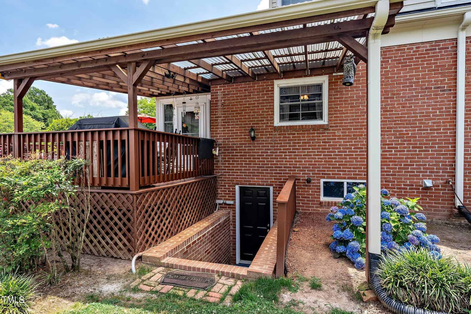 Wooden deck with pergola attached to a red-brick house. Below is a black door leading into a basement. Blue hydrangeas and greenery add color to the yard, and a grill is on the deck under a partly cloudy sky.