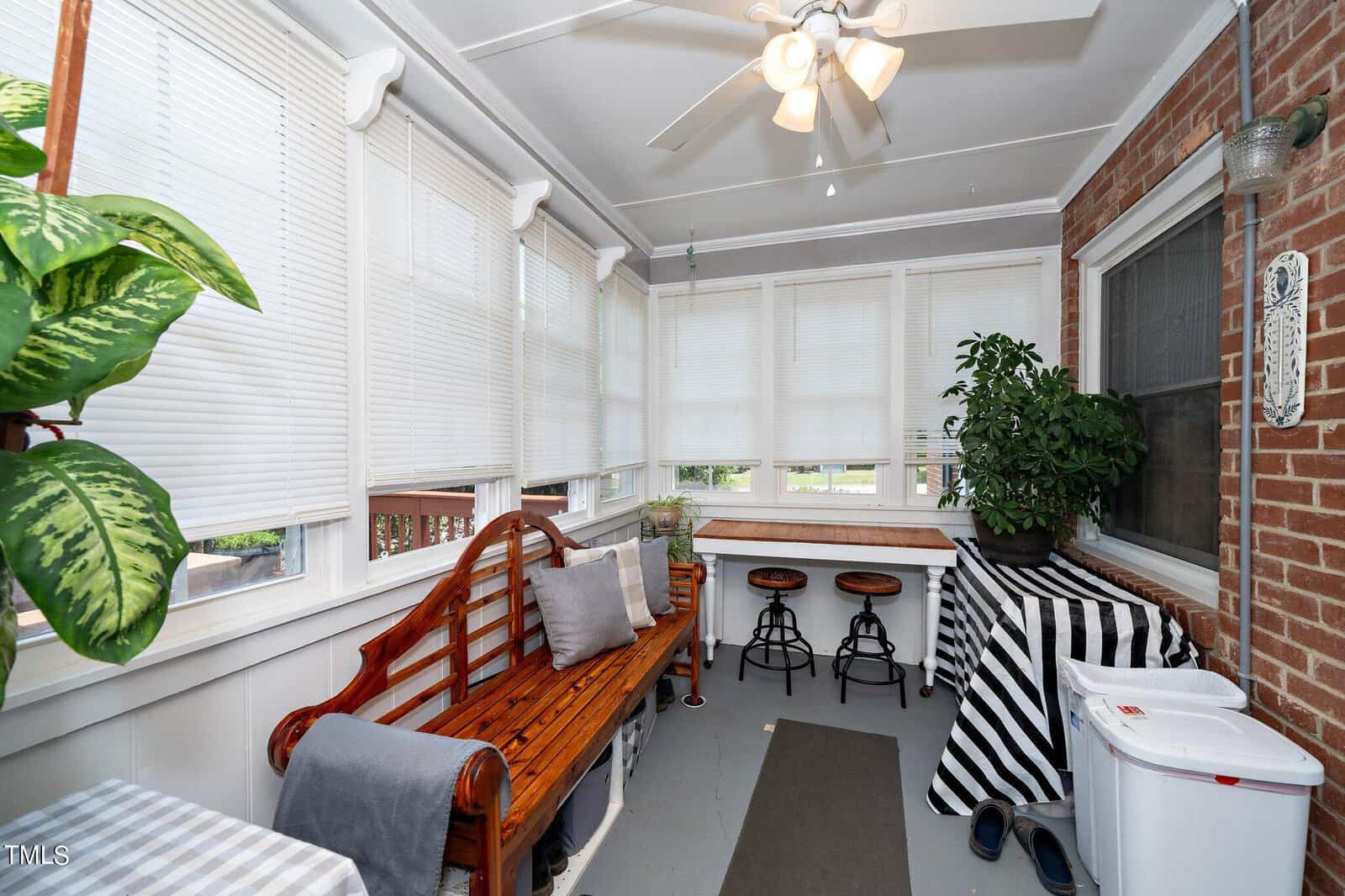 A cozy sunroom with a wooden bench, cushions, and a small table with bar stools. Large windows with blinds let in plenty of light. There's a ceiling fan above and potted plants for decoration. A brick wall adds a rustic touch.