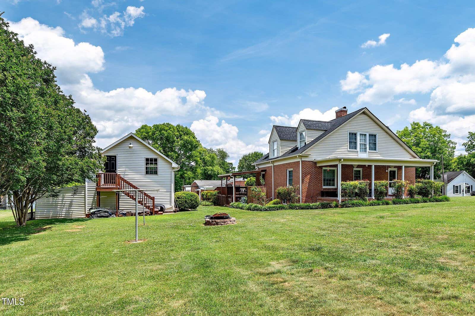 A two-story brick house with a porch, surrounded by green lawn and trees, is pictured on a sunny day. A smaller detached building with an exterior staircase is nearby, alongside a small round fire pit. Fluffy clouds fill the blue sky.