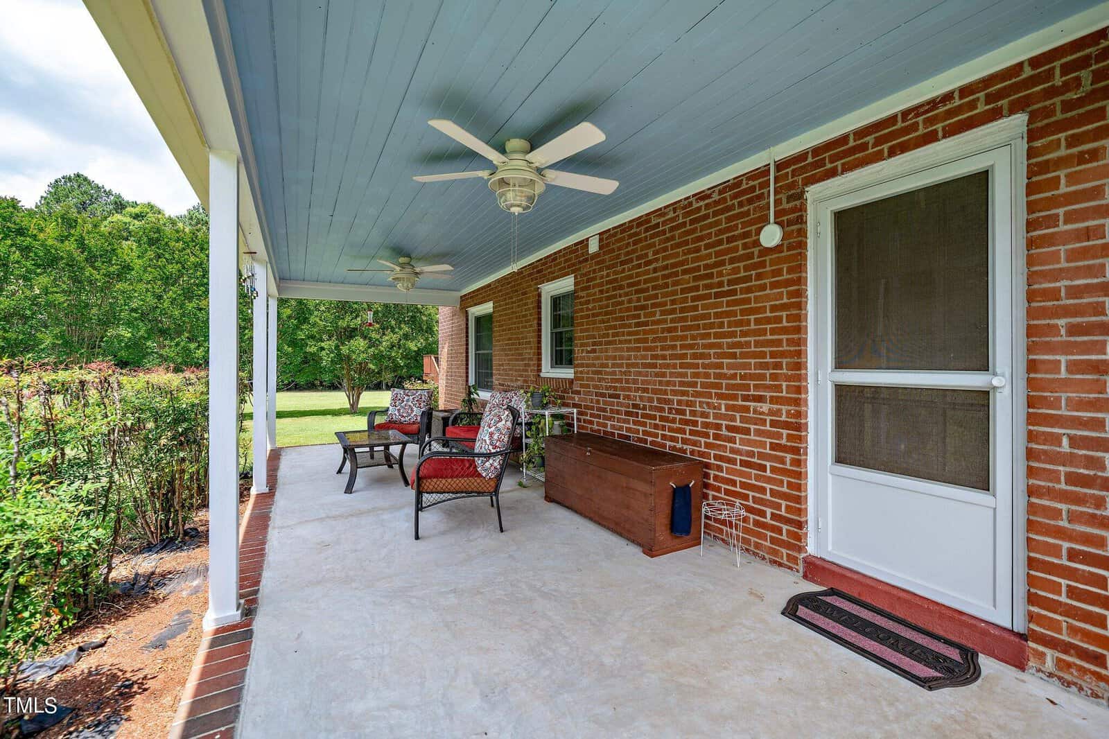 A covered patio with a blue ceiling, featuring a brick wall and a white screen door. Furnished with cushioned chairs and a bench, the area is shaded by two ceiling fans. The surrounding greenery is visible beyond the edge of the patio.