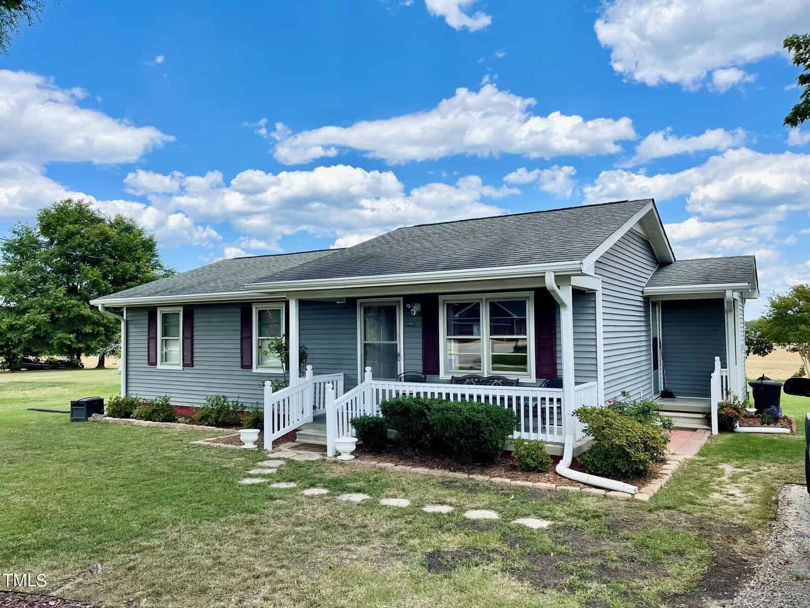 A single-story blue house with white trim, a front porch with a white railing, and a few steps leading up to it. Situated at 2485 Rock Mill Road in Henderson, NC, the yard is grassy with a pathway of stepping stones. The background features a bright blue sky with scattered clouds and some trees.