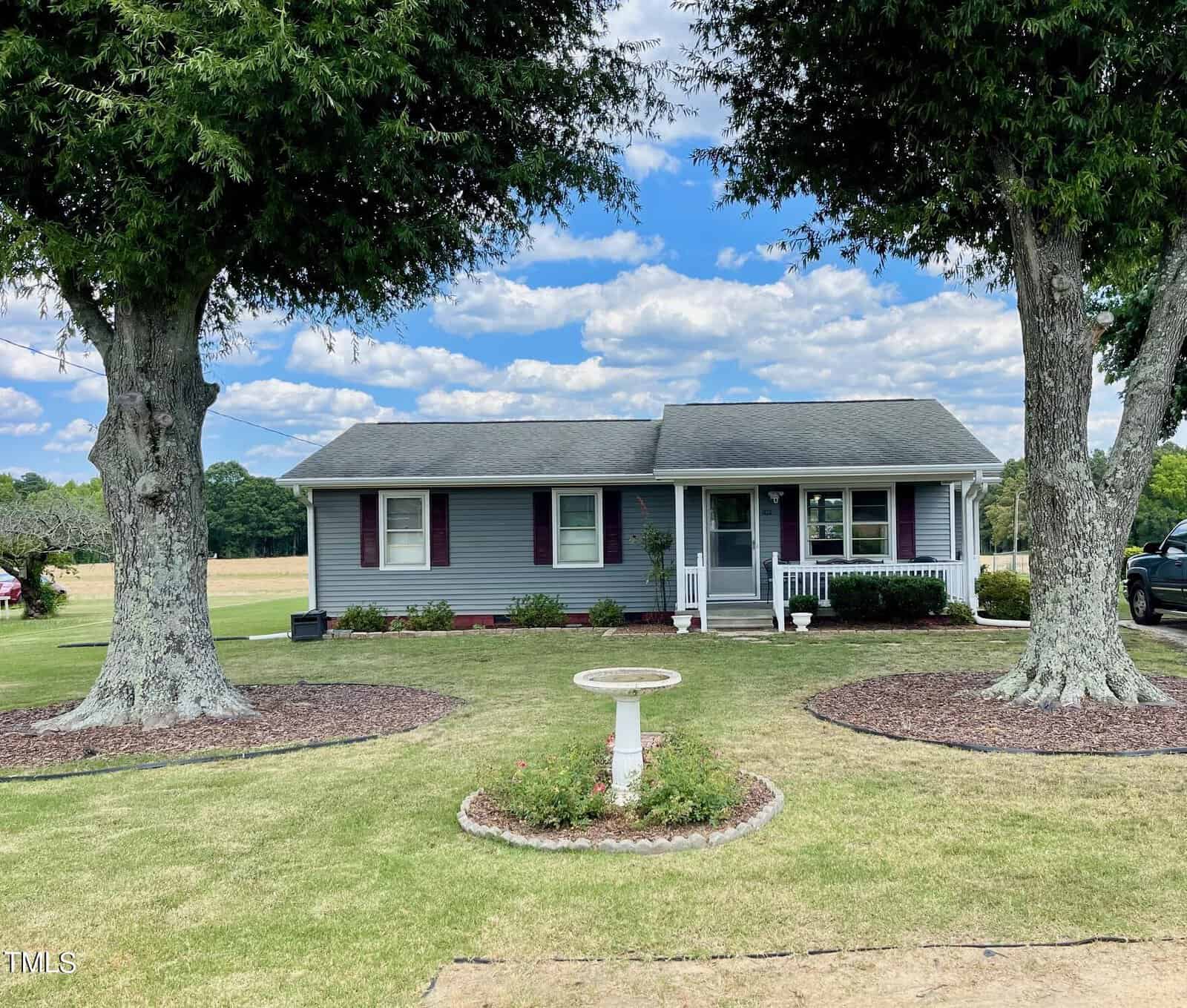 A small, single-story house with gray siding and a front porch. Two large trees frame the house on either side. A birdbath sits in the center of a well-kept lawn, which is surrounded by landscaped flowerbeds. The sky is partly cloudy.