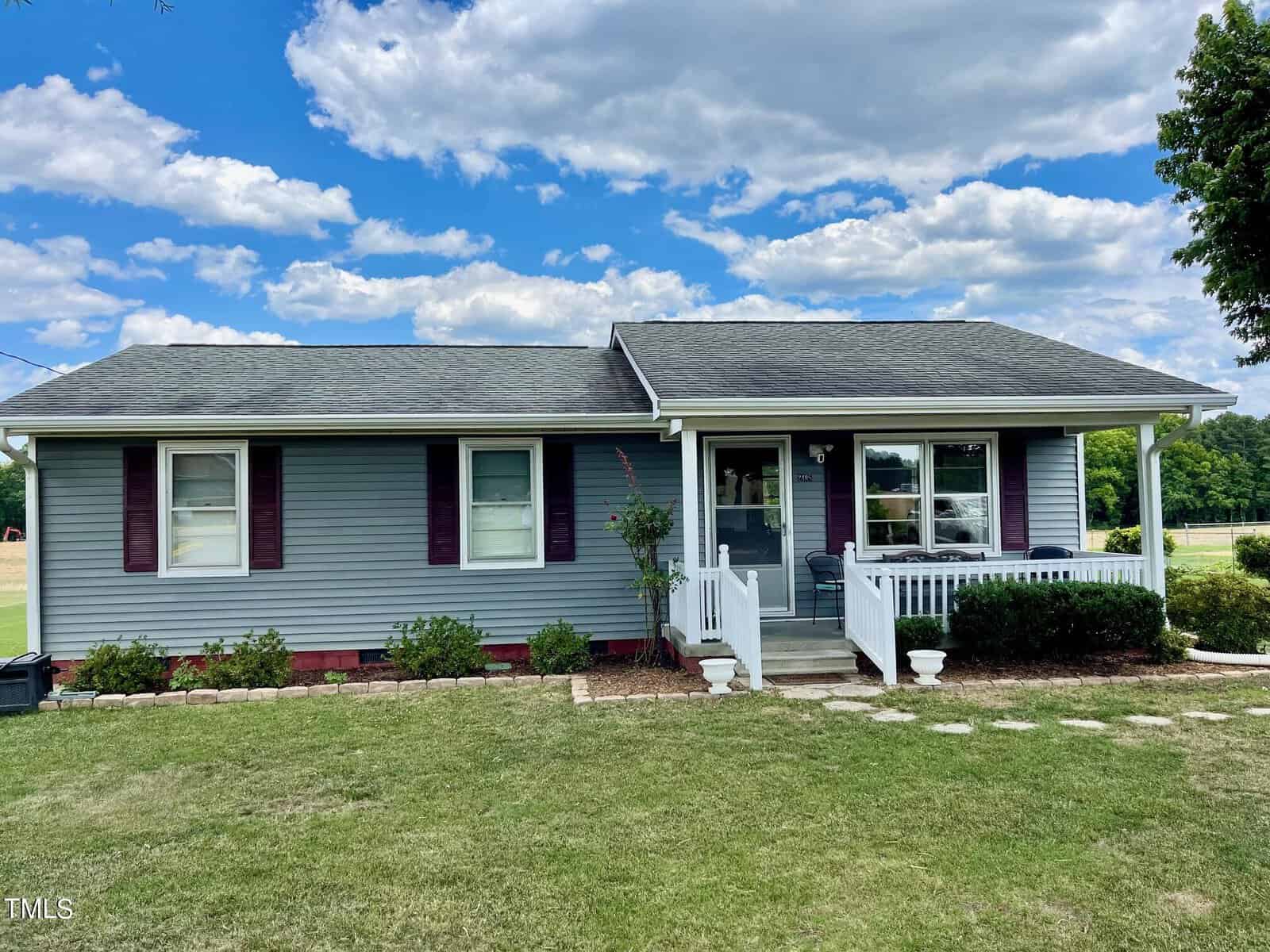 A small, single-story house with gray-blue siding, white trim, and a dark roof. It features a front porch with white railings, red shutters on the windows, and a well-maintained lawn with a stone pathway leading to the porch. There are trees and a field in the background.