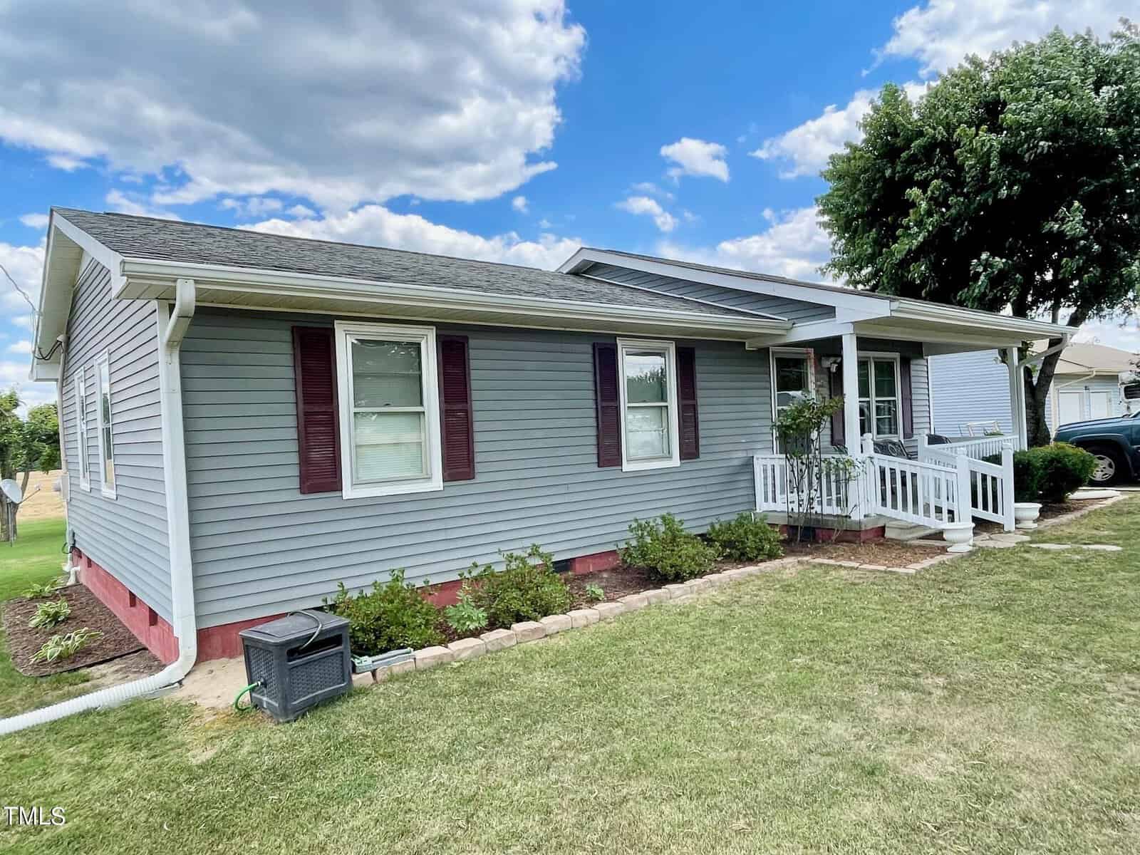 A single-story house with gray siding and red shutters. It features a small front porch with white railings, a well-kept lawn, and shrubs lining the foundation. The sky is partly cloudy, and there are trees in the background.
