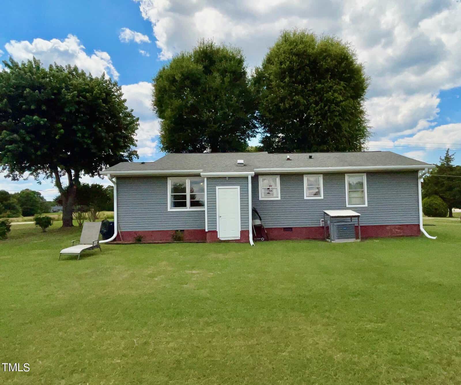 A small, single-story gray house with a red brick foundation and white trim. The house has a grassy front yard with two large trees behind it and a lounge chair on the lawn. The sky is partly cloudy. The front door is white, and there are several windows.