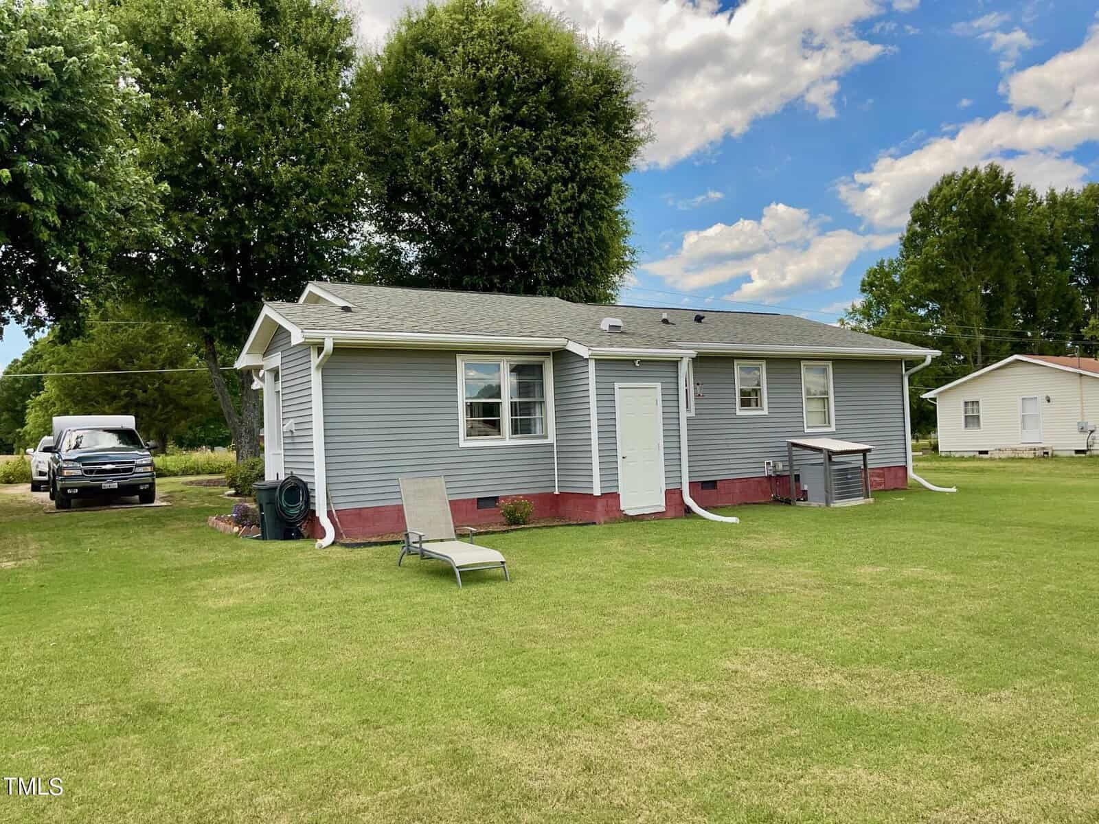 A single-story house with gray siding and red brick foundation sits on a well-maintained lawn. The house has a white door on the side and multiple windows. A lounge chair is placed on the grass, and a tree and a white pickup truck are visible in the background.