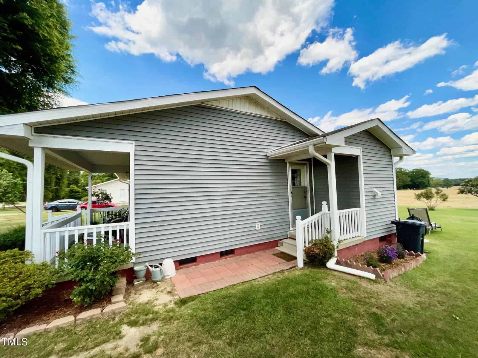 A small, single-story house with gray siding and white trim. It features a small porch with a white railing, a green lawn, and a blue sky with scattered clouds. There are shrubs and plants along the brick foundation.