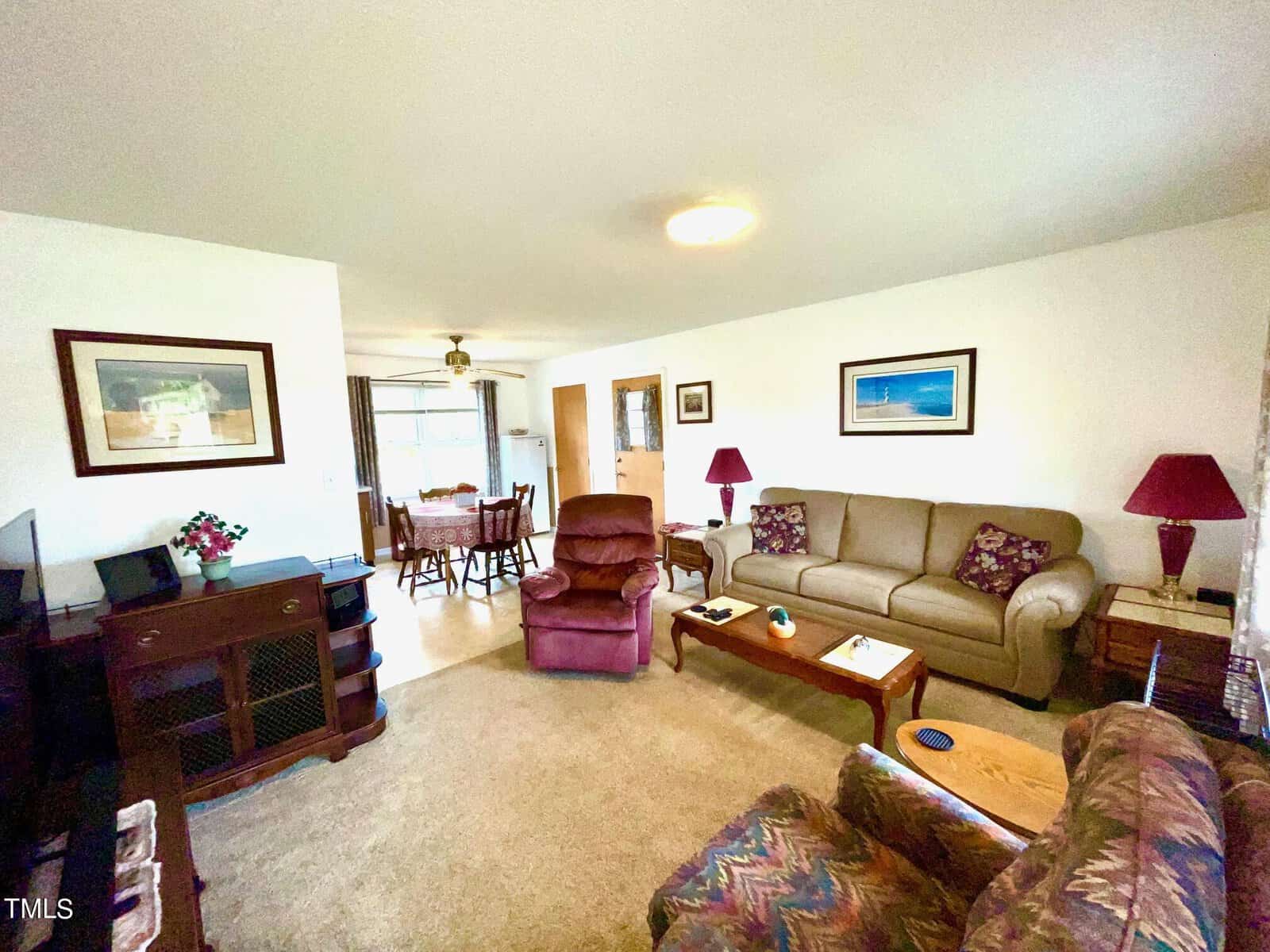 A cozy living room with sofas, an armchair, and a recliner around a coffee table. The room features framed pictures on the walls, a TV, a lamp, and a side table with a vase of flowers. The adjacent dining area is partially visible. The room is well-lit and carpeted.