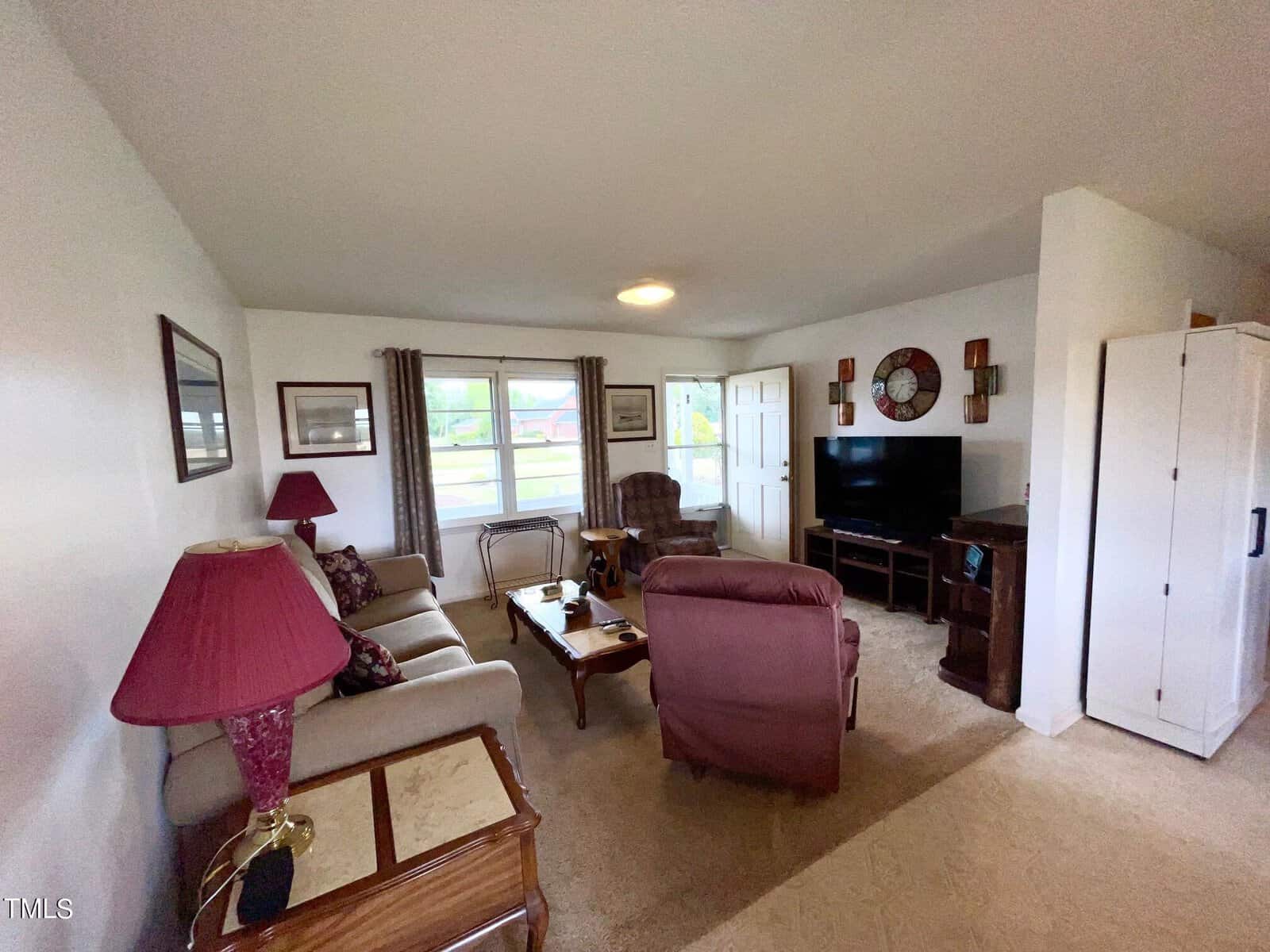 A cozy living room with beige walls and carpet, featuring a sofa, two armchairs, a wooden coffee table, and a flat-screen TV on a stand. The room is decorated with framed pictures, a purple lamp, and a decorative wall clock. A window lets in natural light.
