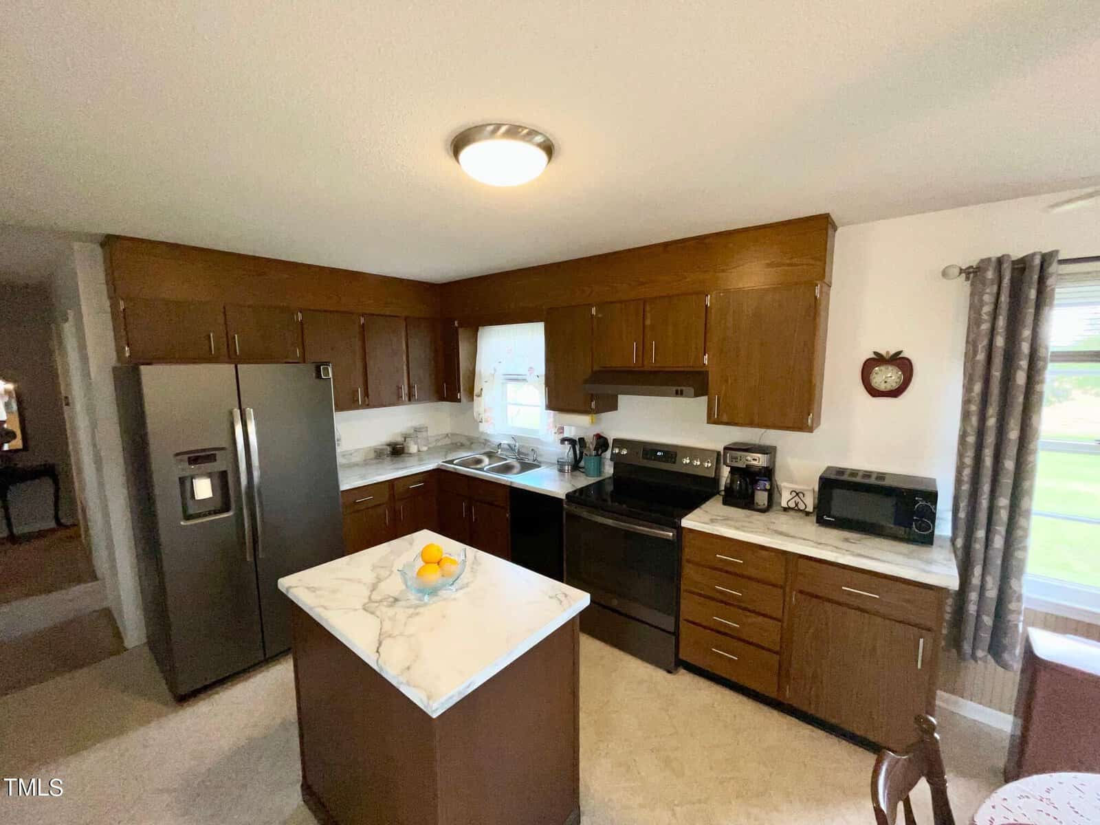 A kitchen with wooden cabinets, a marble countertop island, stainless steel appliances including a refrigerator, oven, and dishwasher. A bowl of fruit is on the island. The window to the right overlooks a grassy yard. A small dining table is partially visible.