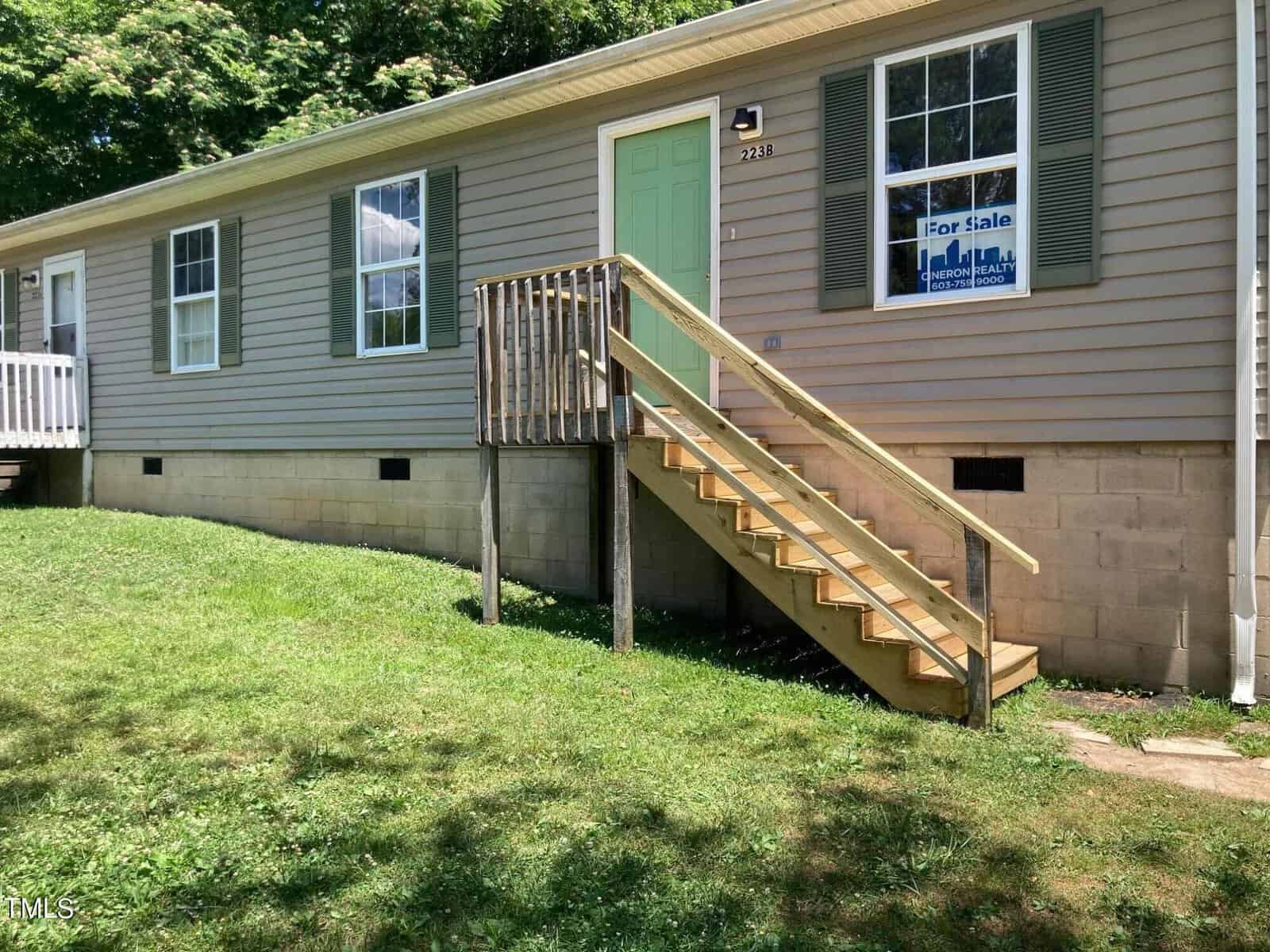 Nestled at 223 Maple Drive in charming Oxford, this single-story house boasts light gray siding and green shutters. A quaint wooden staircase leads to a welcoming green front door. The sunny yard is lush and grassy, with a "For Sale" sign prominently displayed in the window.