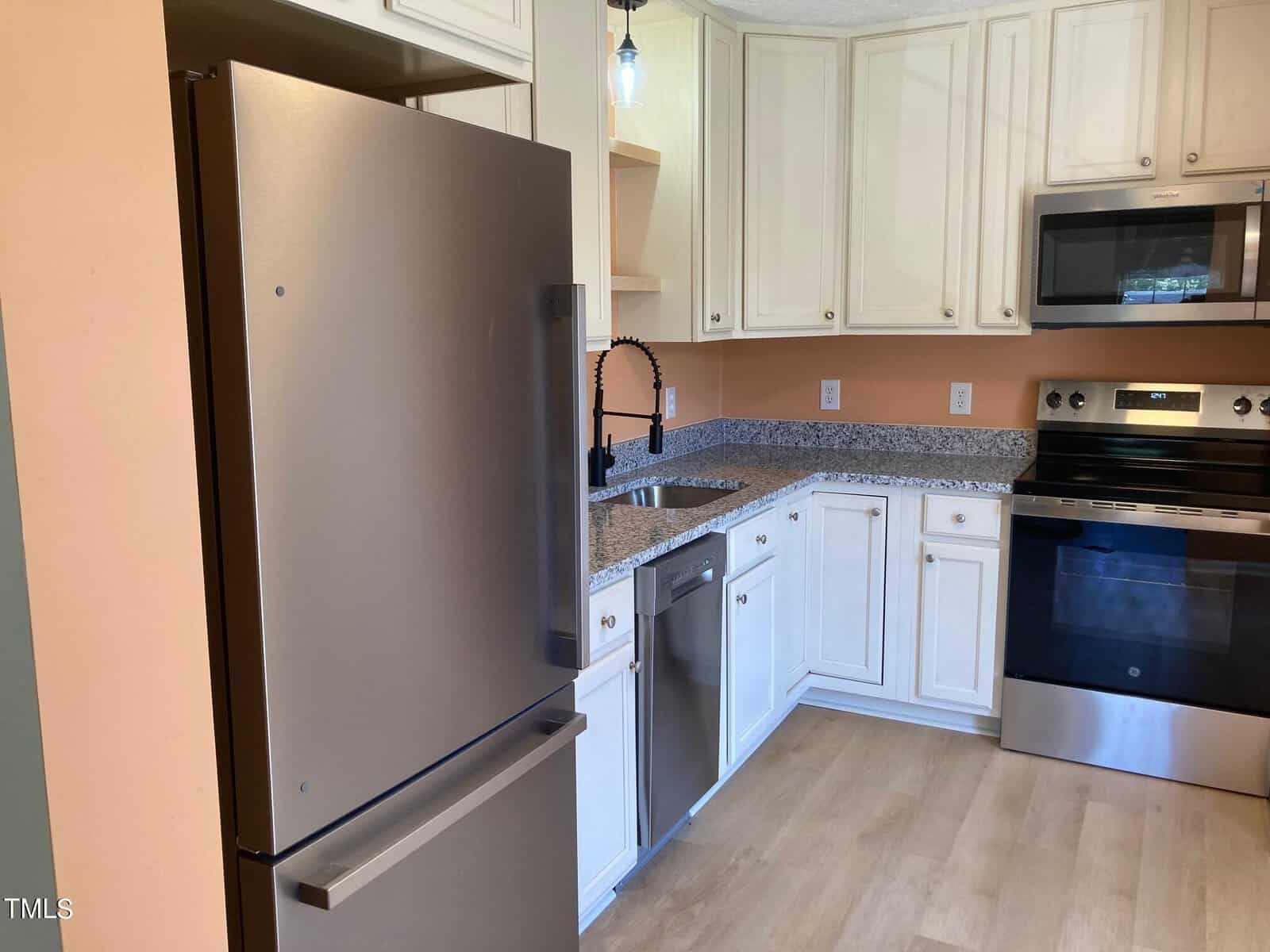 A modern kitchen with stainless steel appliances, including a refrigerator, oven, and dishwasher. White cabinets contrast with granite countertops. A black faucet is installed over a sink, and the floor is wooden. The lighting fixture is visible above.